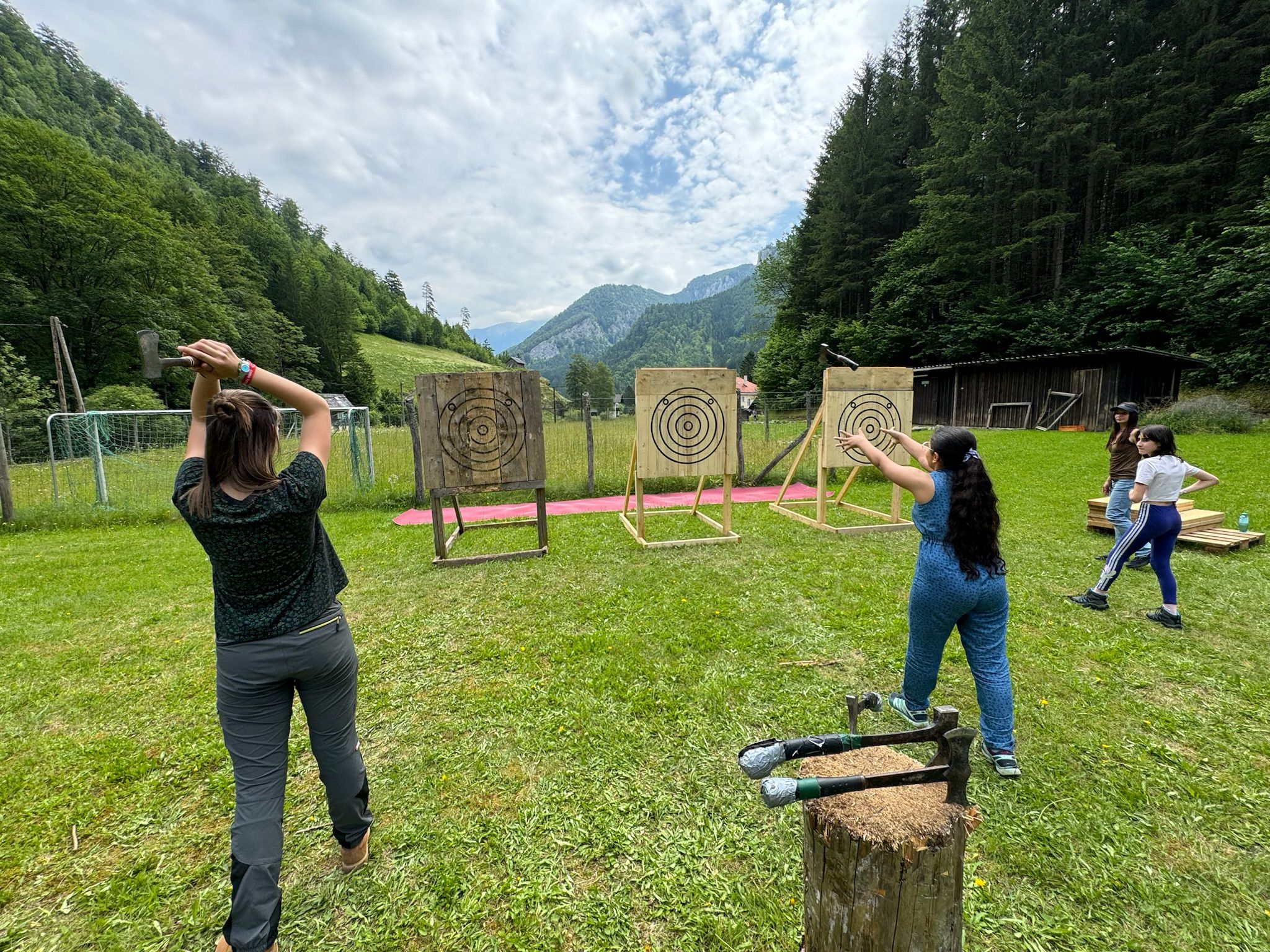 People throw axes at outdoor targets surrounded by forest and mountains.