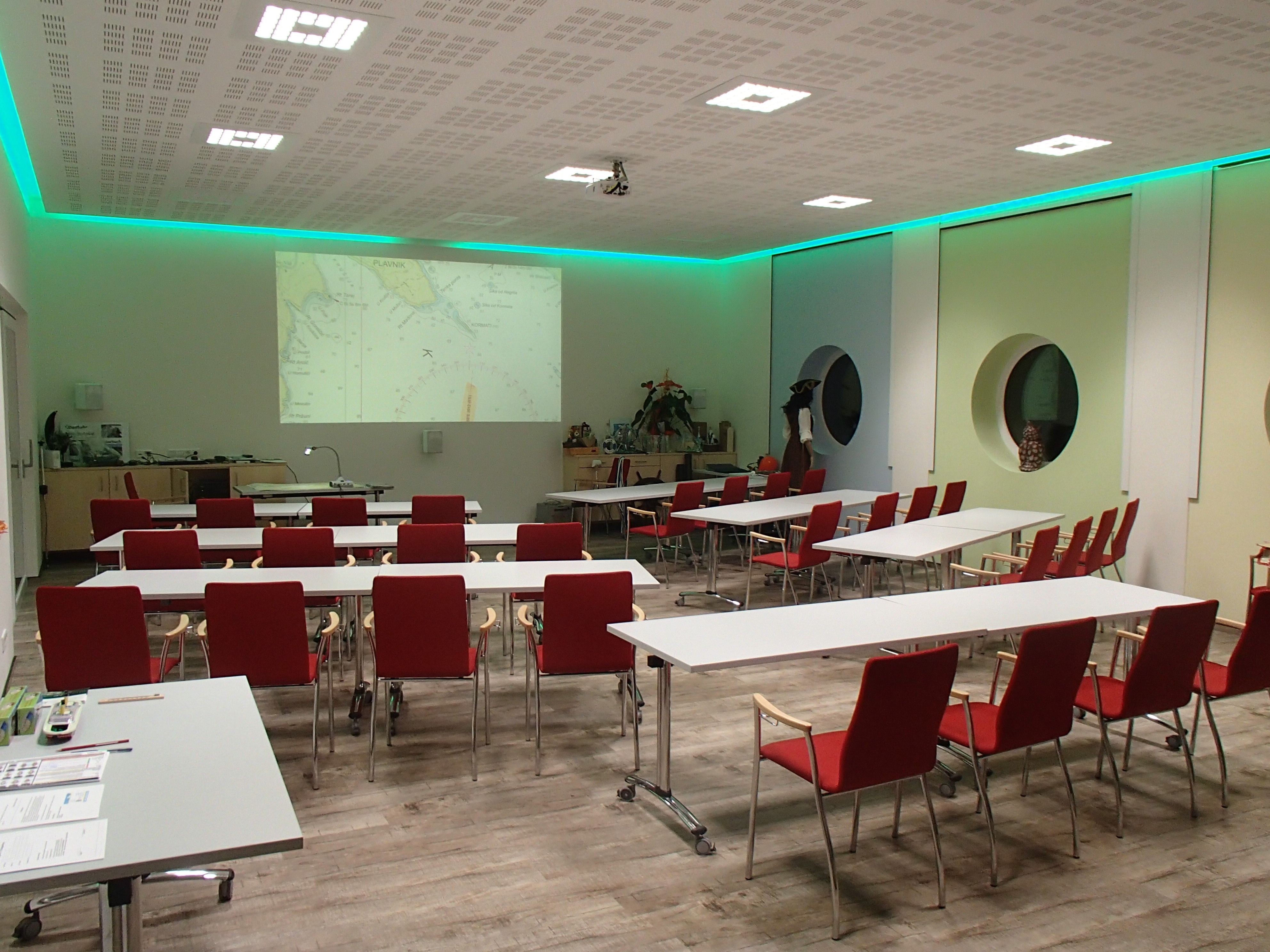 Training room with red chairs and white tables, projector shows nautical chart on the wall.