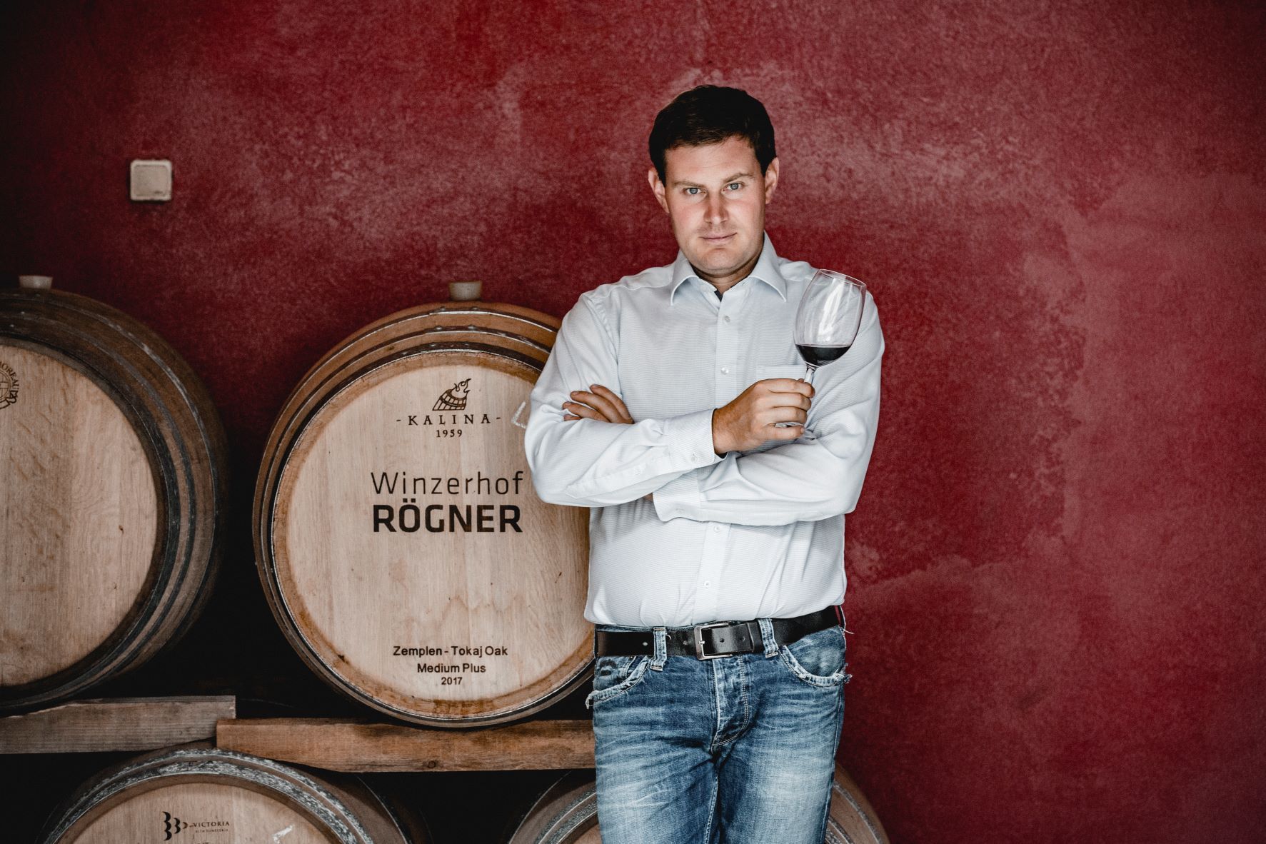 A man stands in front of wine barrels and holds a glass of red wine in his hand.