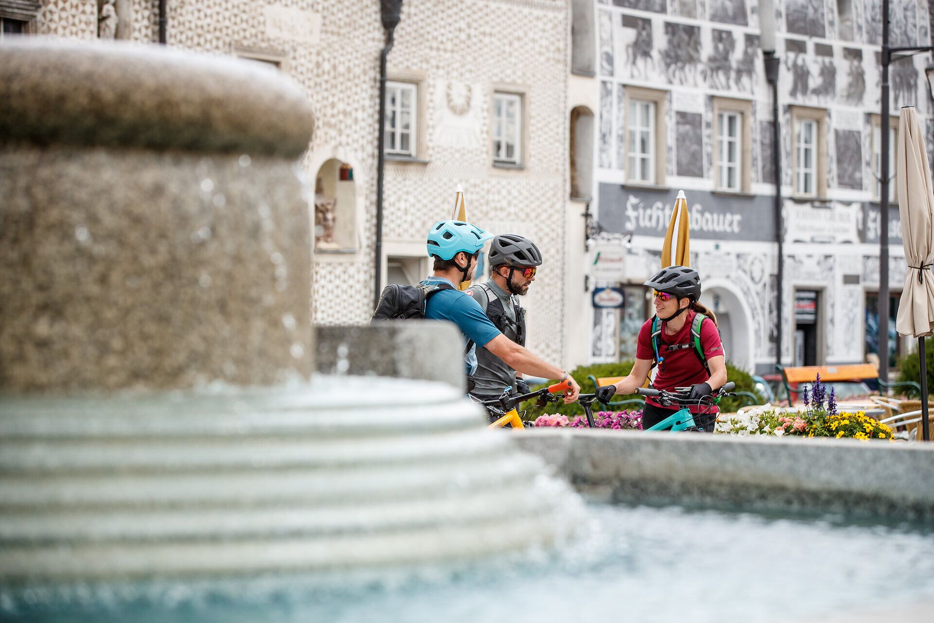 Three cyclists enjoy a relaxing break in the town square, surrounded by blooming flowers and charming architecture. The atmosphere is lively and inviting, while the fresh air and nature invite you to explore.