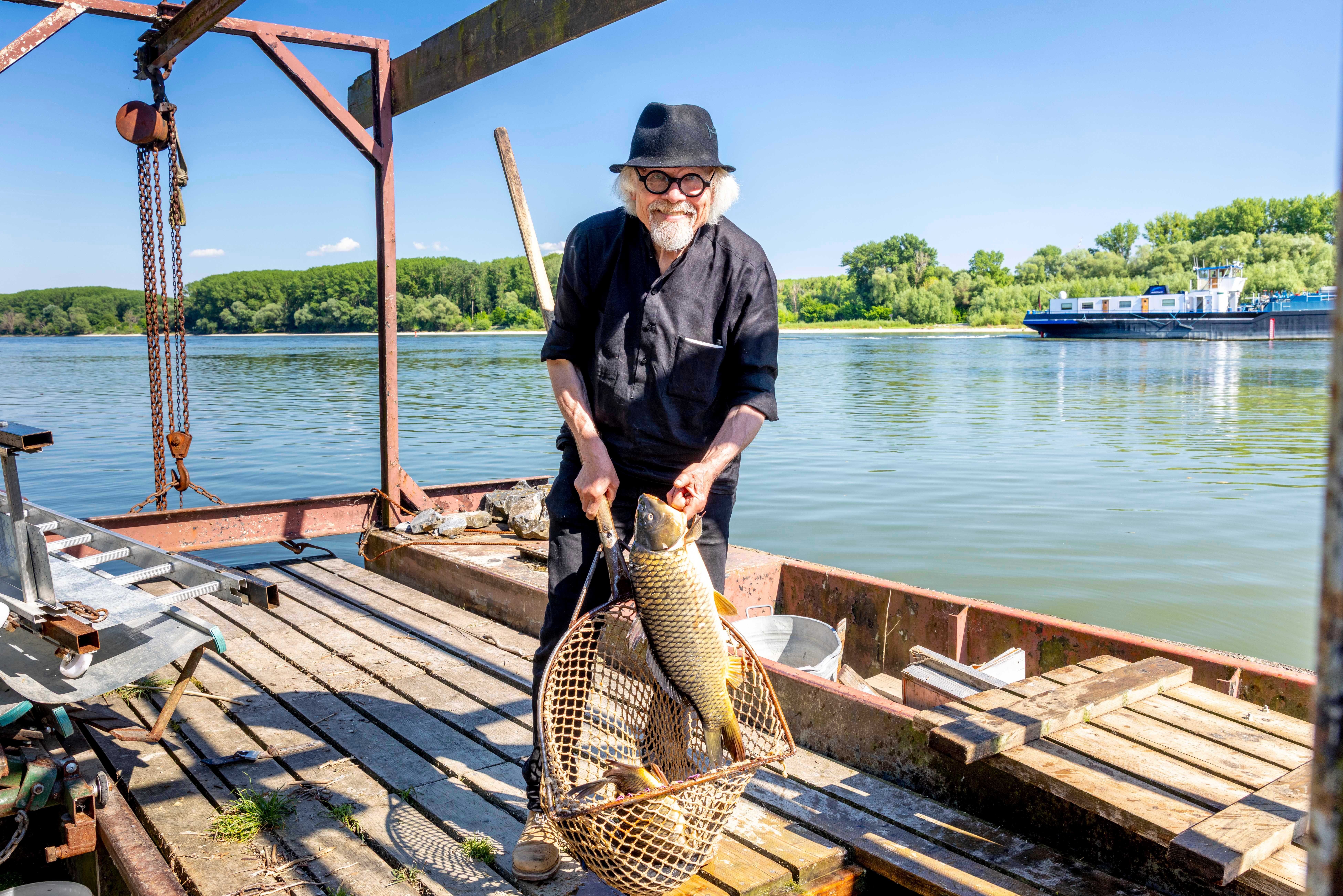 A man with a hat holds a large fish in a net on a wooden bridge on the river (Danube).