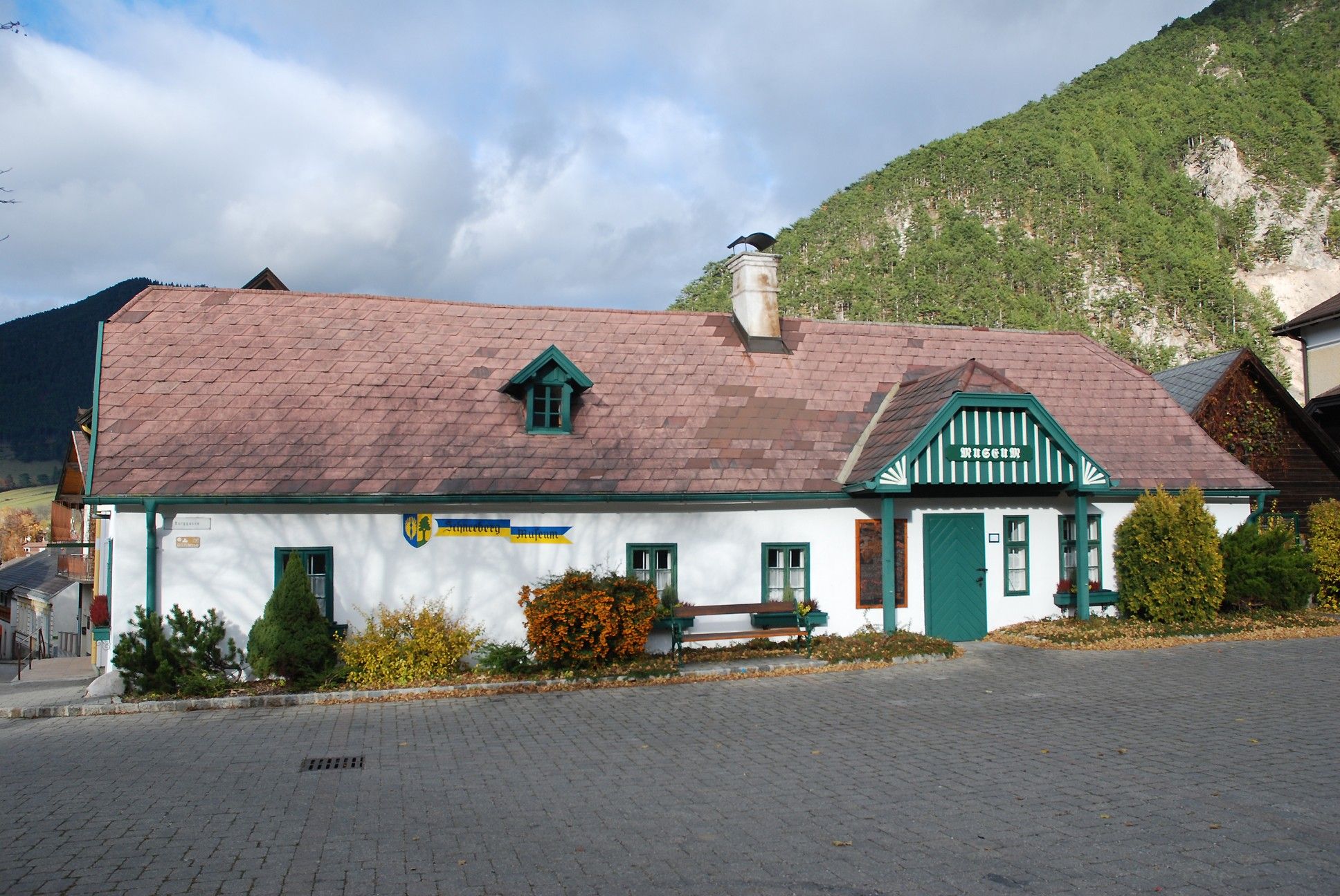 Schneeberg Museum Puchberg, a traditional building with a green roof and chimney, surrounded by mountains and trees.