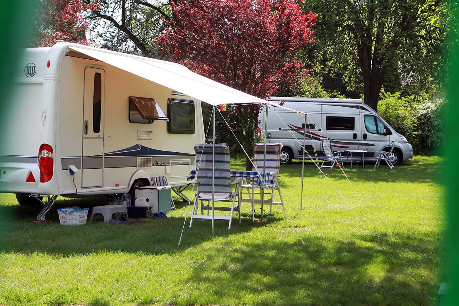 Caravan and motorhome on a green campsite with chairs and table under an awning.
