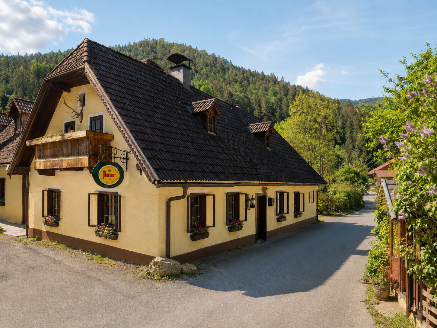 A traditional inn with a yellow facade and a dark shingle roof, set in a rural setting with a forest in the background.