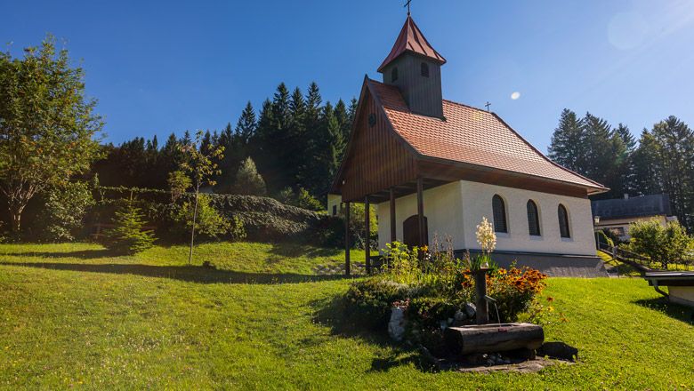 Small chapel with a red roof on a green meadow, surrounded by trees and flowers.
