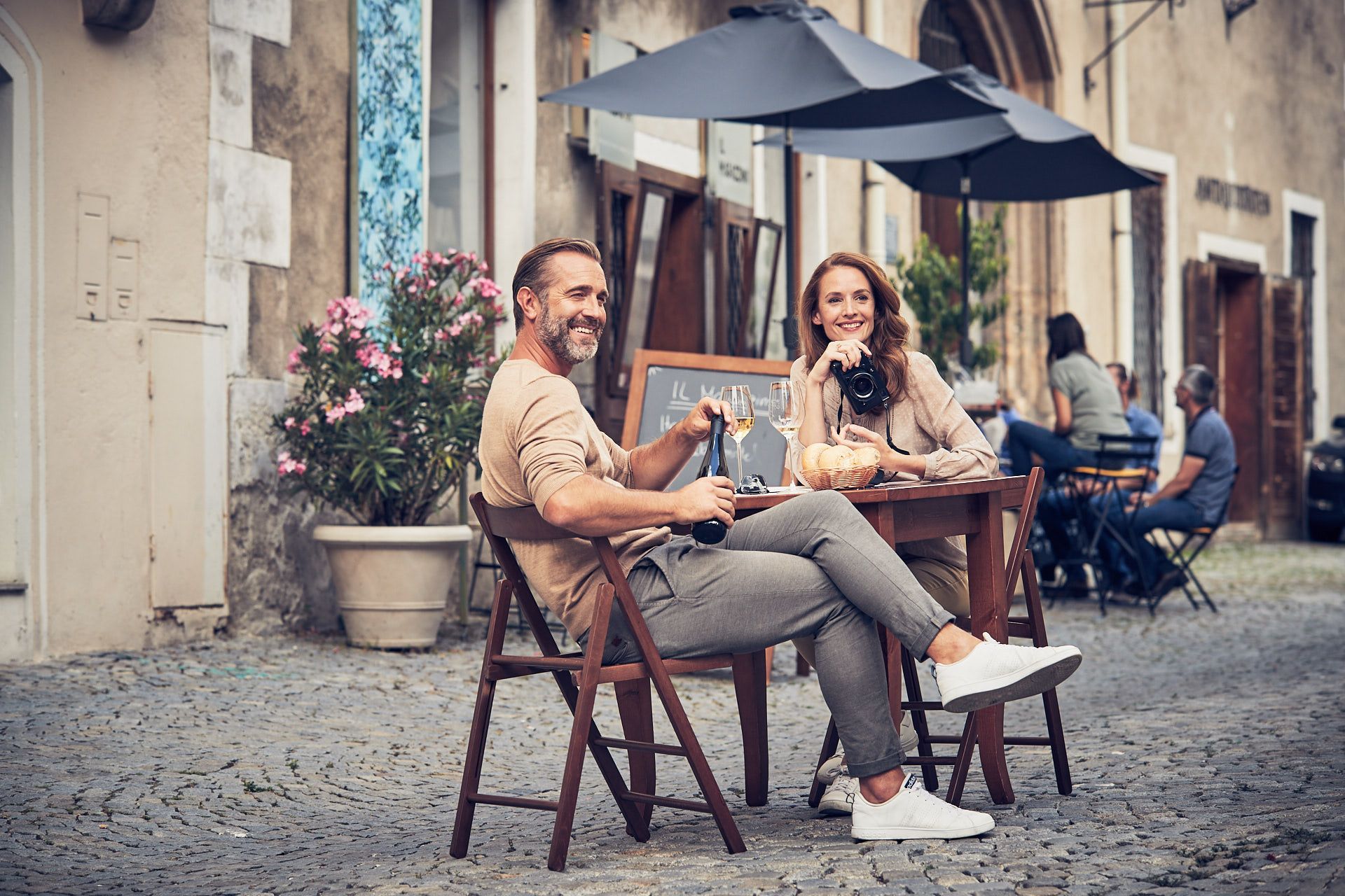 A couple sits at a table in the old town of Krems, drinking wine and smiling. Other guests and historic buildings can be seen in the background.