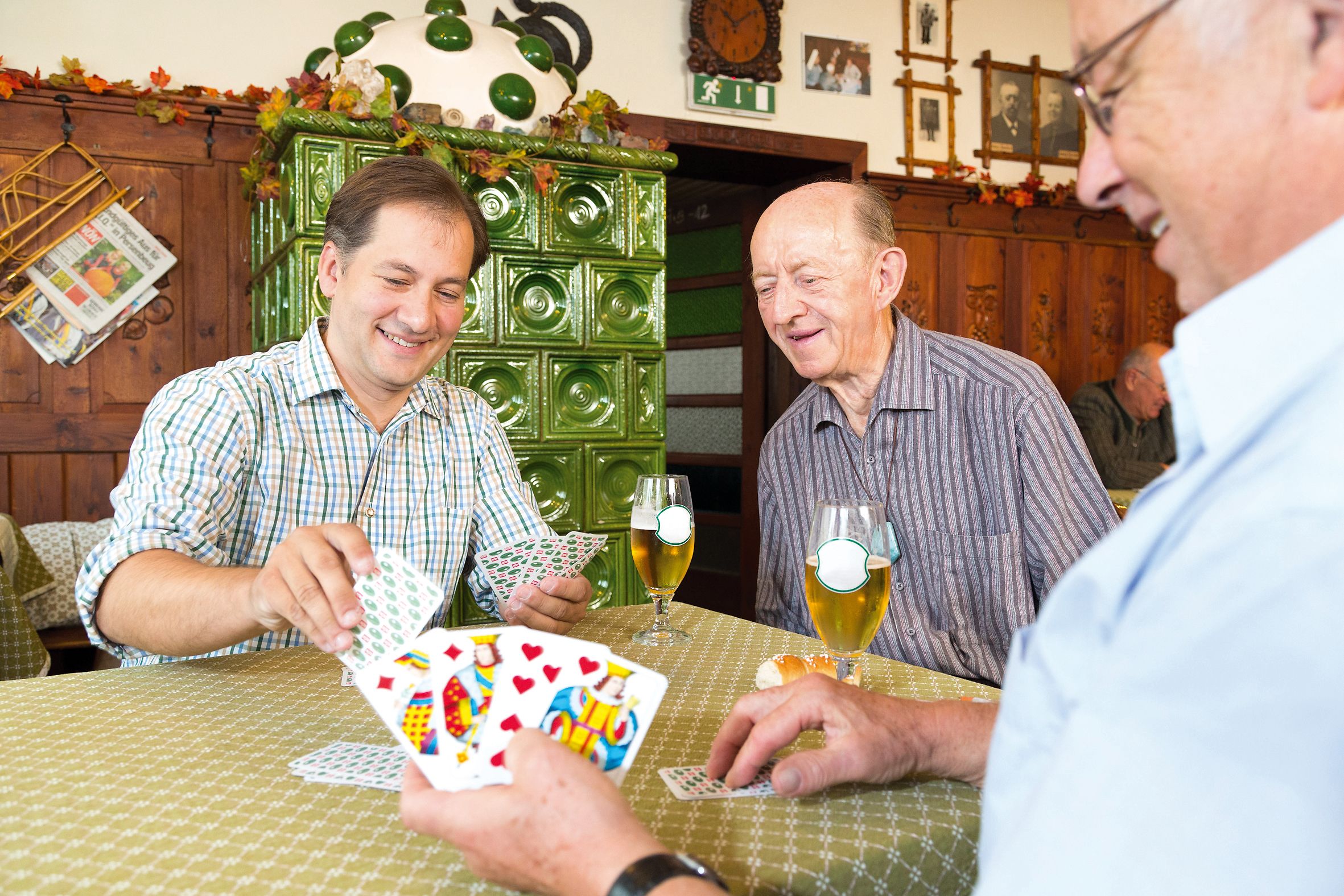 Three men playing cards in an inn with a green tiled stove in the background.