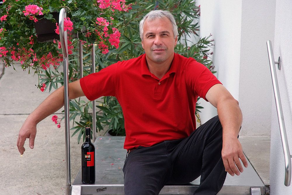 A man in a red shirt sits on a staircase next to a bottle of wine and flowers.