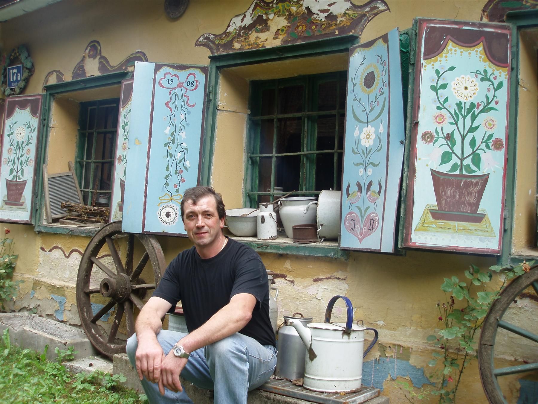 Man sitting in front of an old building with painted shutters and old household objects.