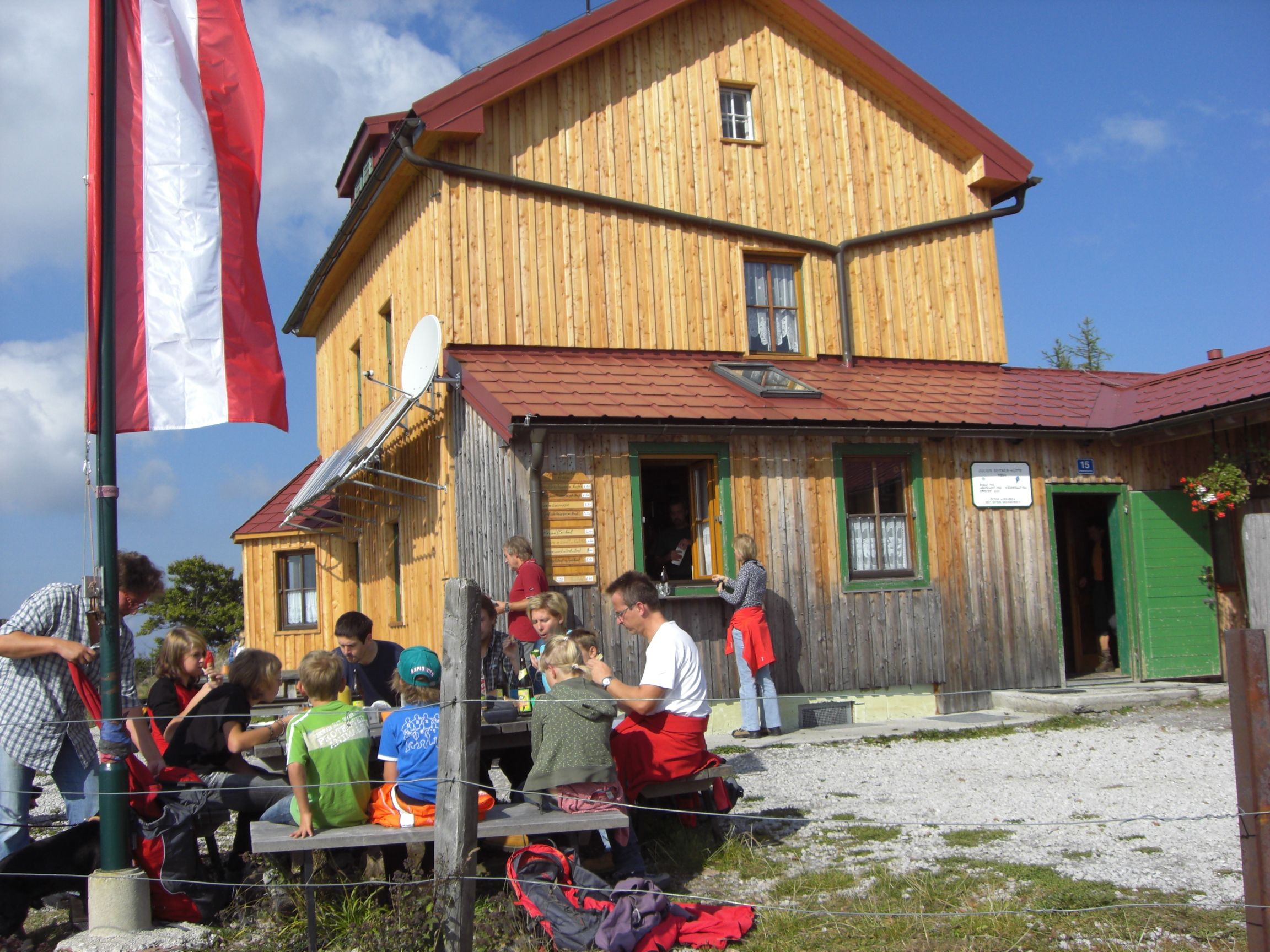 A group of people sit in front of a mountain hut with a wooden façade and Austrian flag.