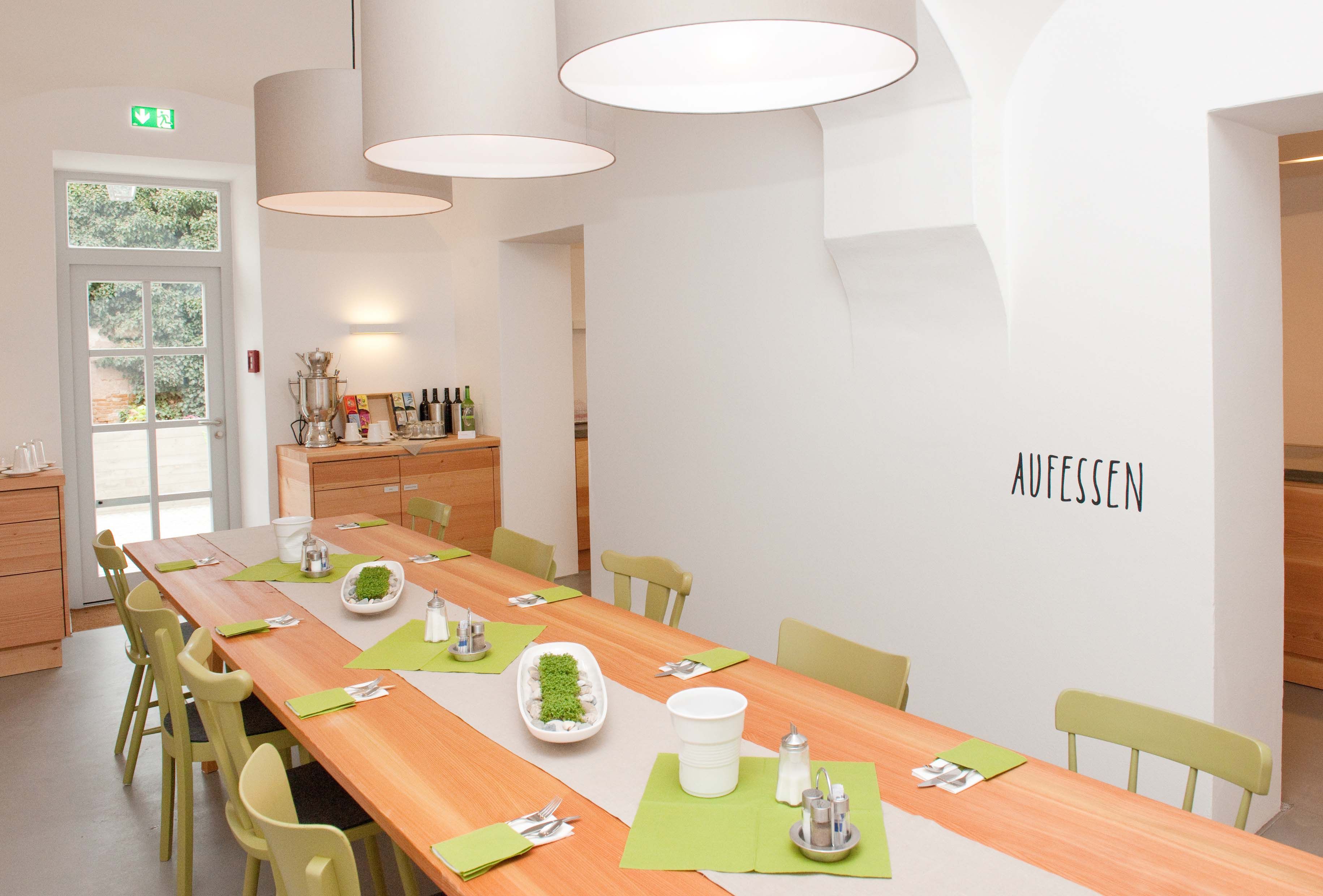 A bright dining room with a long wooden table, green chairs and decorations. 'AUFESSEN' is written on the wall.