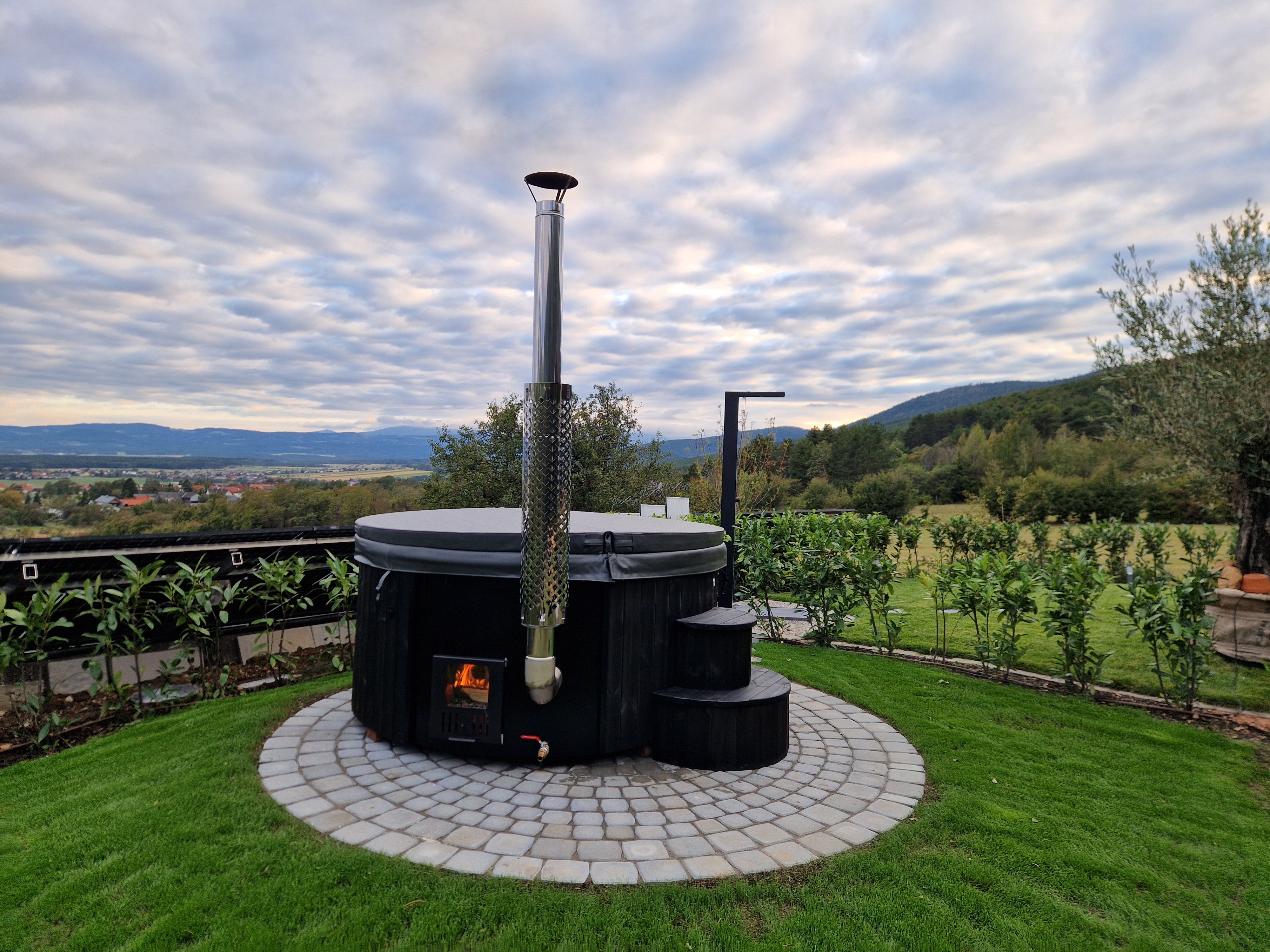 A round hot tub with an outdoor fireplace, surrounded by green countryside and cloudy skies.