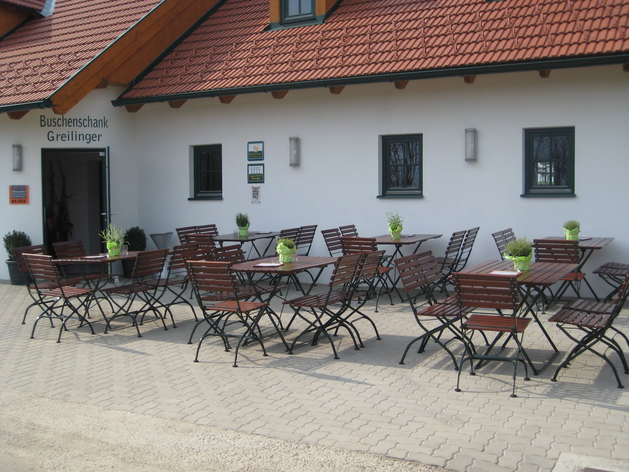 Outdoor area of a Buschenschank (typical tavern) with empty wooden tables and chairs in front of a building with a red tiled roof.