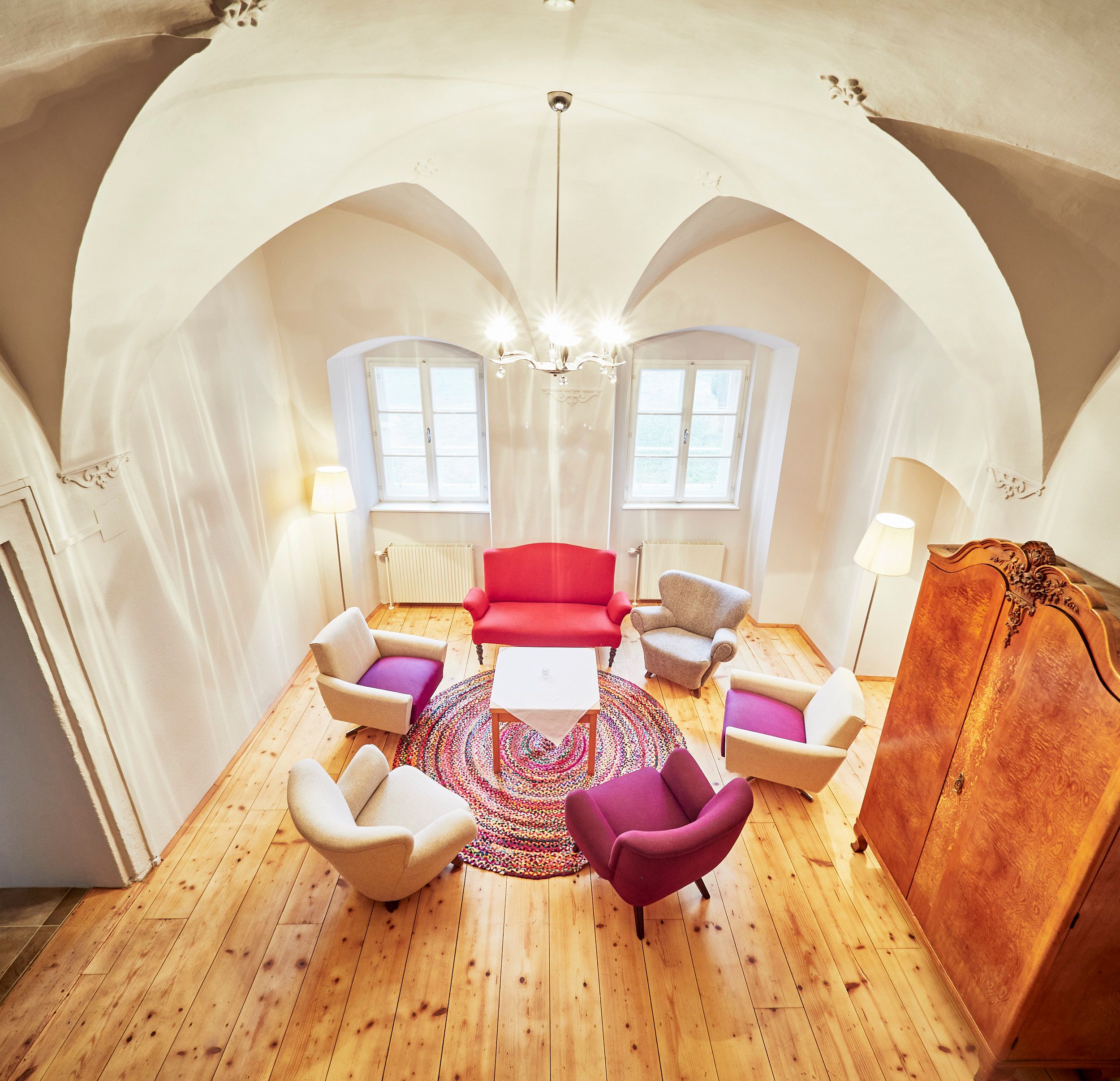 A cozy tea room with colorful armchairs, a red sofa and a round carpet on a wooden floor. A chandelier hangs from the vaulted ceiling.