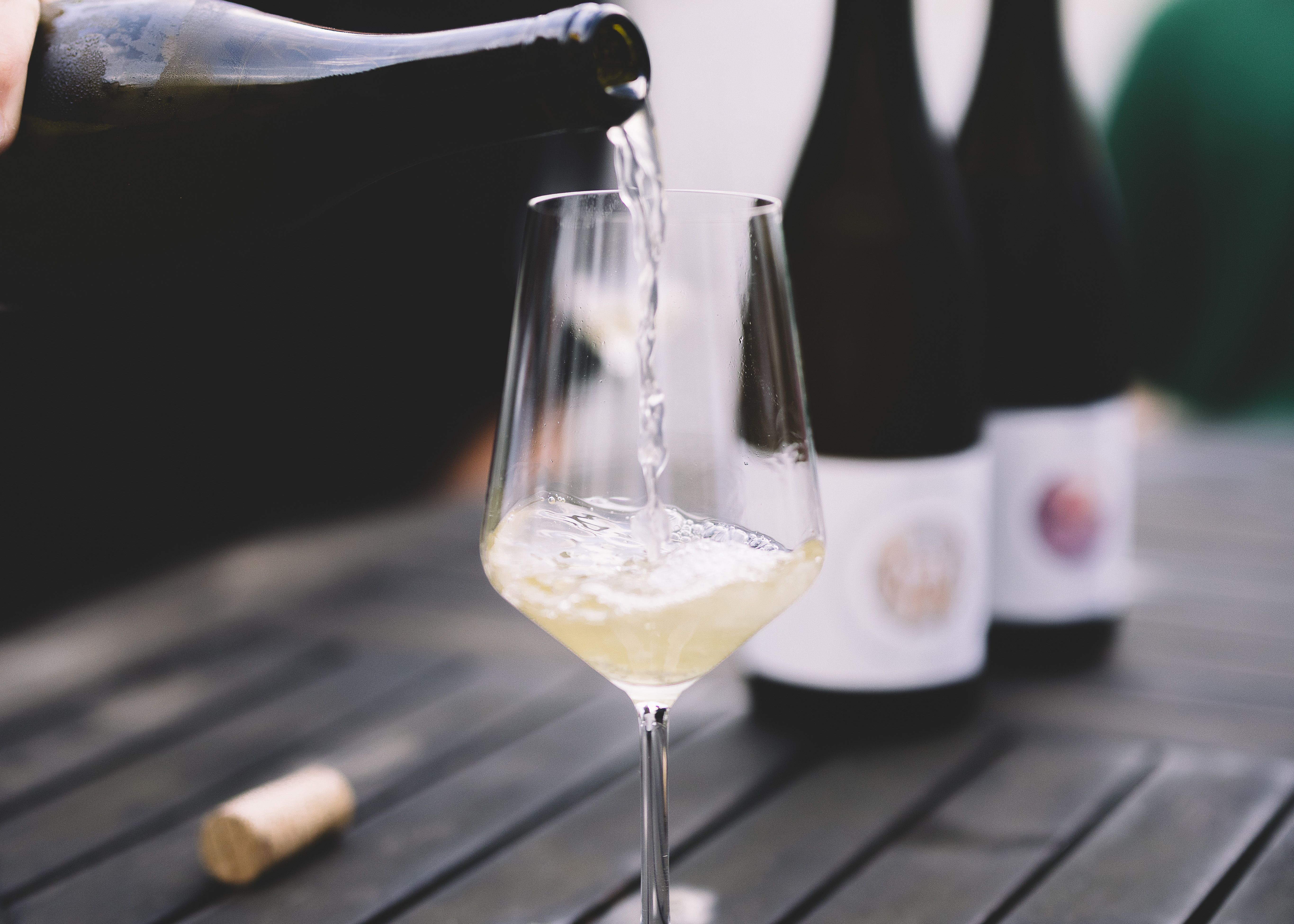 A glass of white wine is poured on a table, with two bottles of wine in the background.
