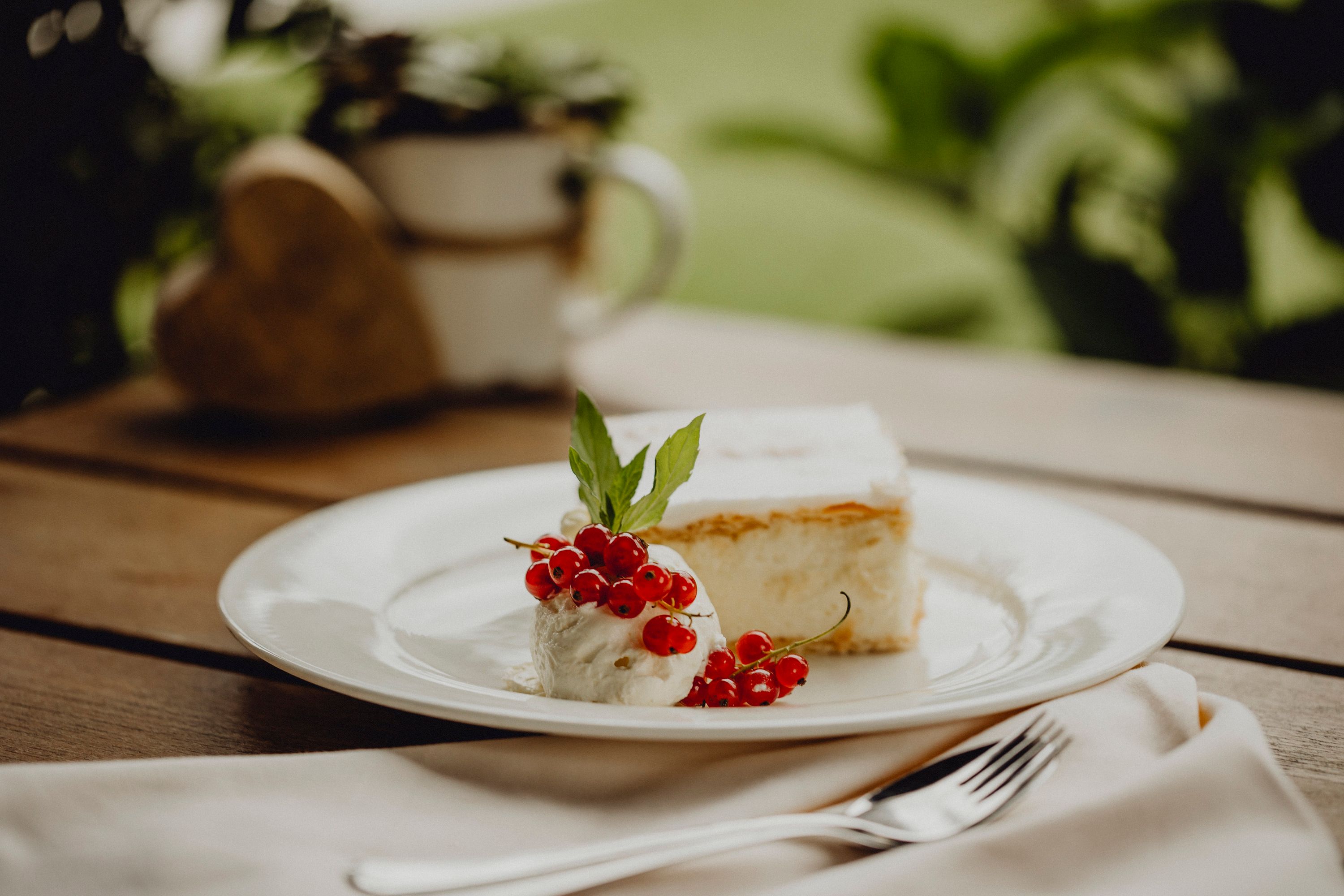 A plate with a cream slice, cream, red currants and a mint leaf on a wooden table.