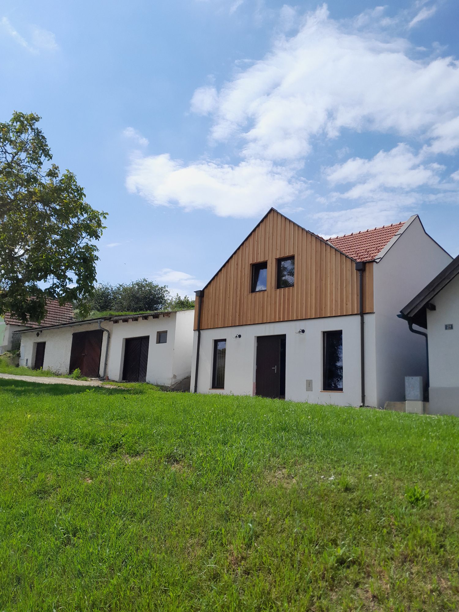 A modern building with a wooden façade and red roof tiles stands on a green hill under a blue sky.