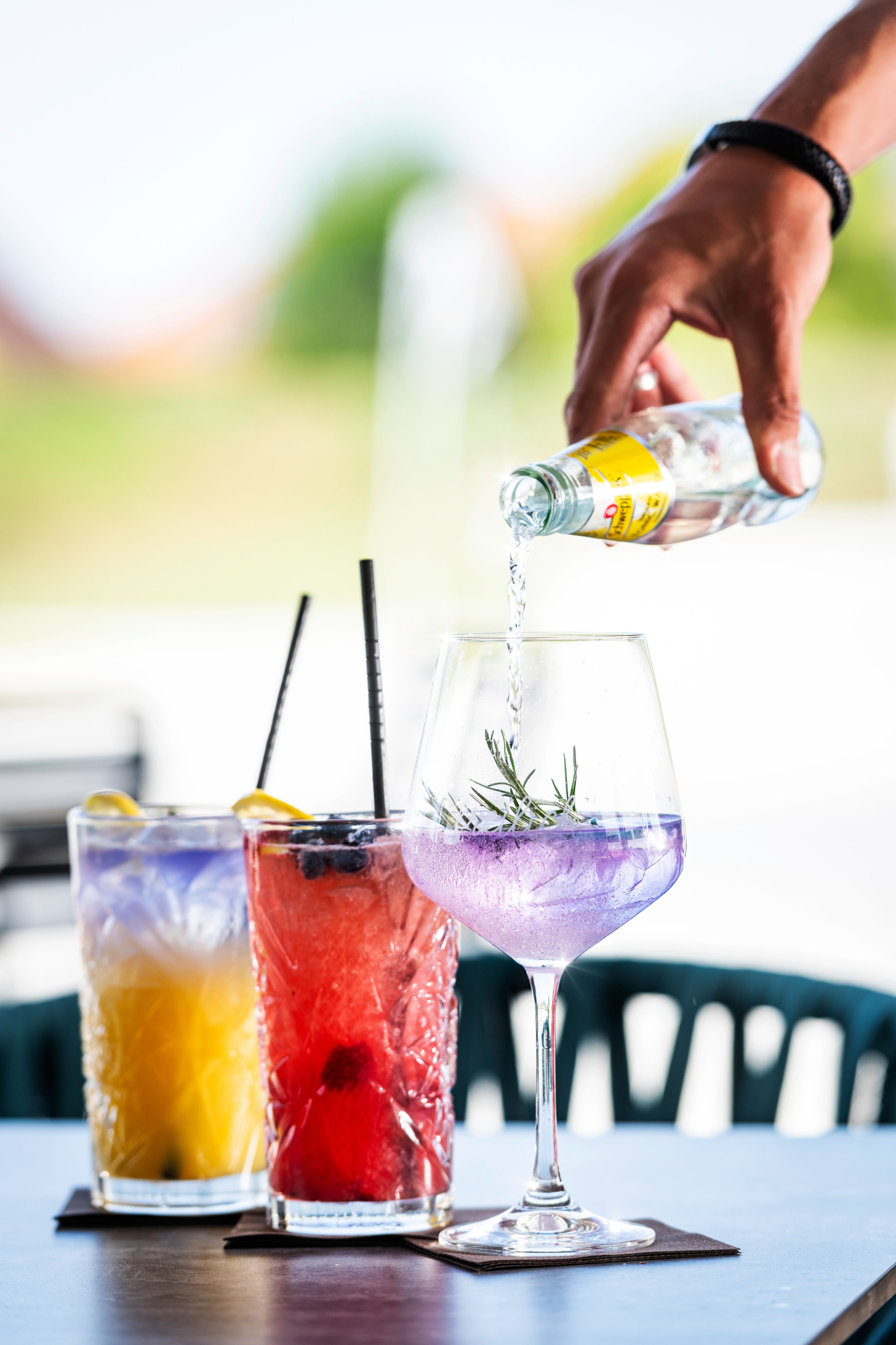 Three colorful cocktails on a table, a hand pours liquid into a glass.
