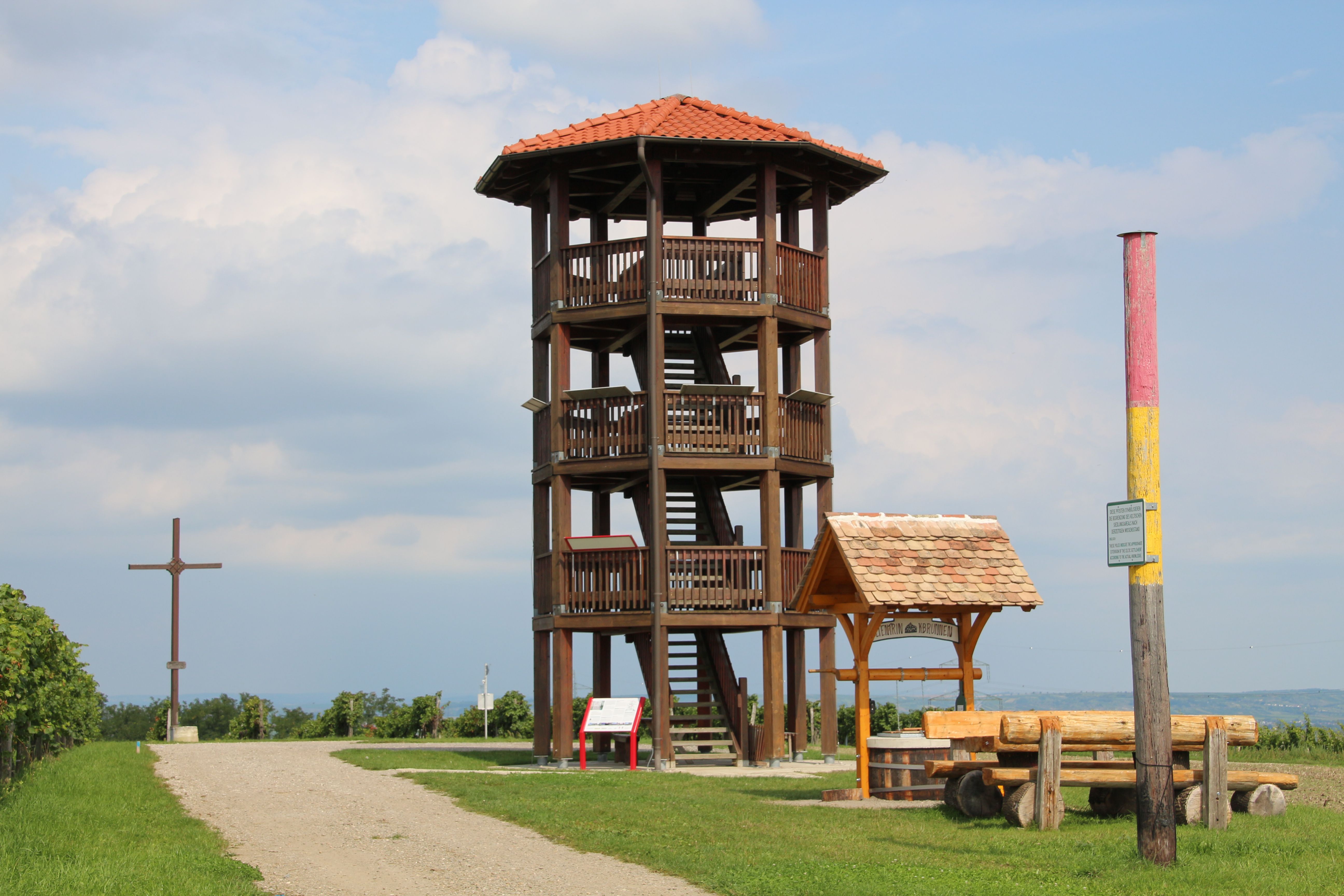 Wooden lookout tower with red roof on a meadow, next to it a wooden cross and a bench.