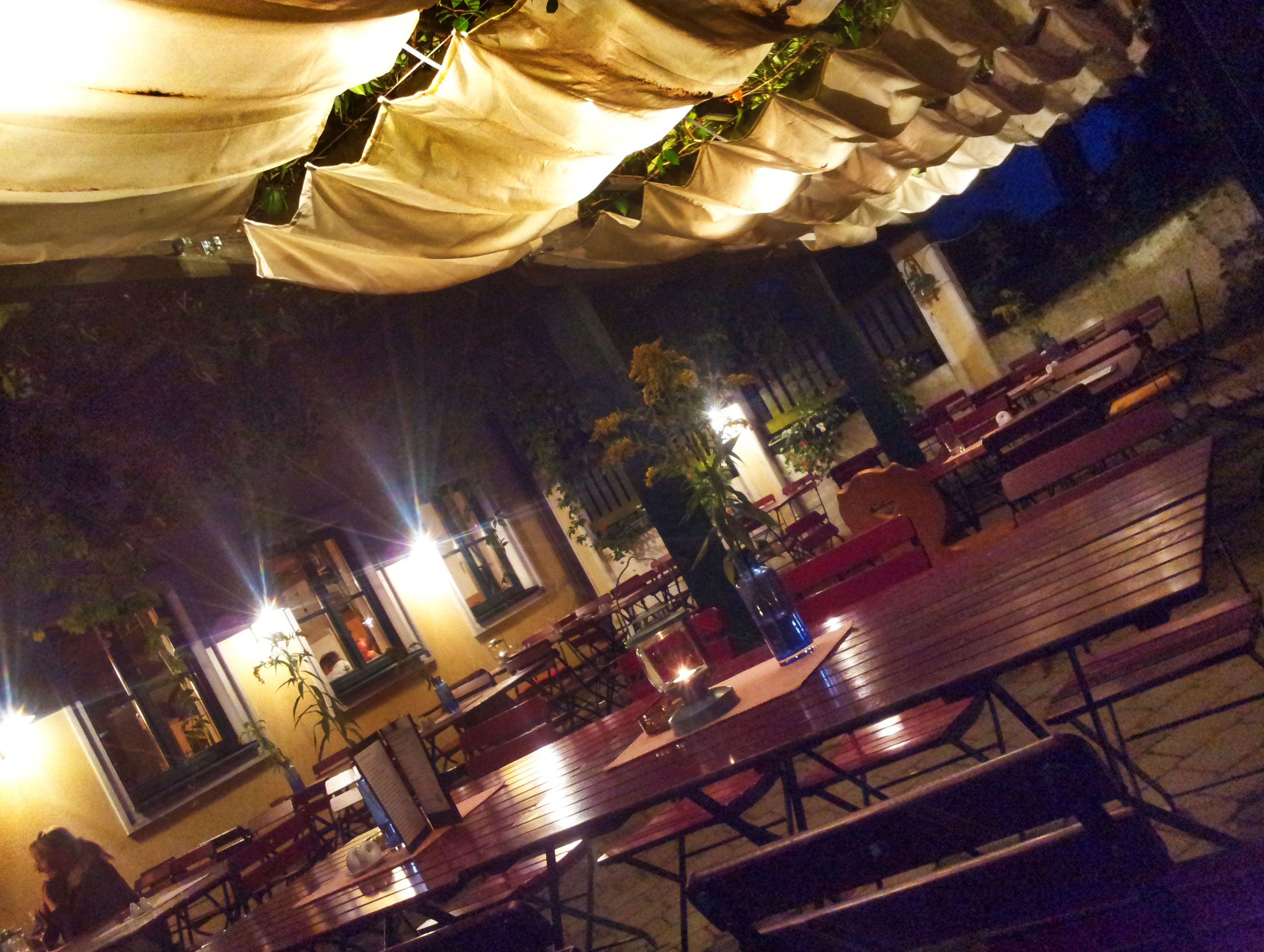 Illuminated terrace with wooden tables and chairs, covered with fabric panels, at night.