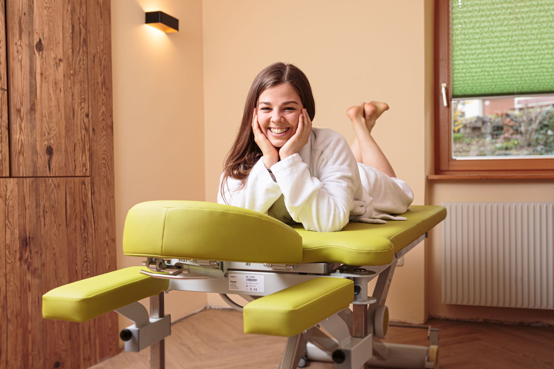 Woman in a bathrobe lying smiling on a green massage table in a wellness room.