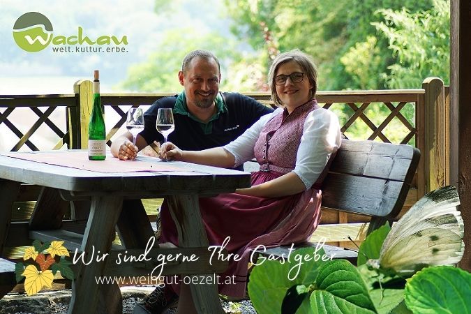 A couple sits at a wooden table outdoors with wine and glasses. Nature can be seen in the background. Text: "We are happy to be your hosts".