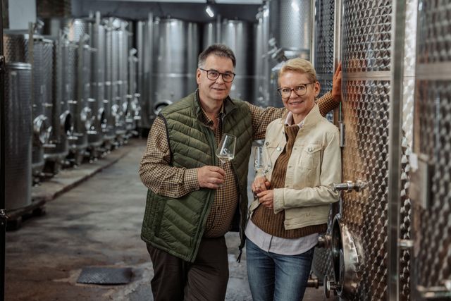 A man and a woman stand in front of large stainless steel tanks in a winery, both holding a wine glass.