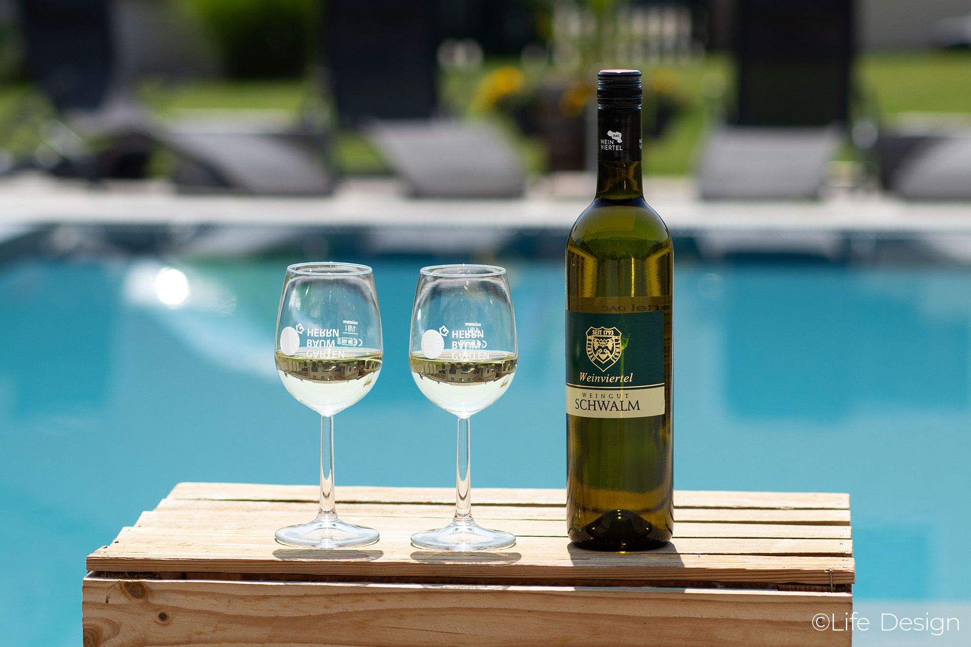 A bottle of wine and two wine glasses on a wooden table by the pool.
