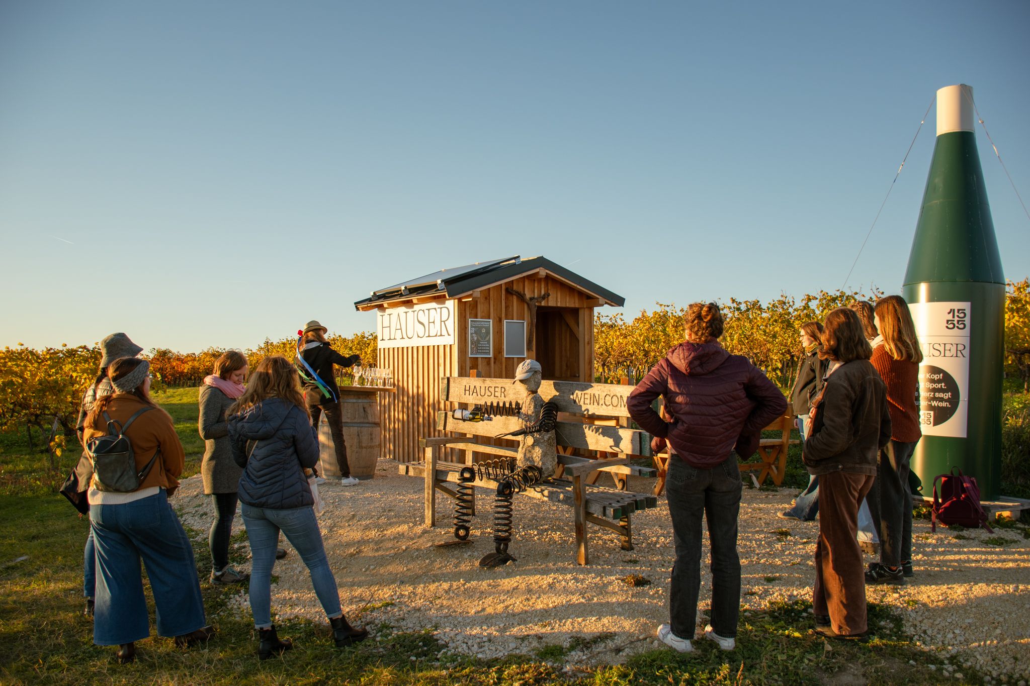 Group of people standing in front of a small wooden house in a vineyard at sunset.