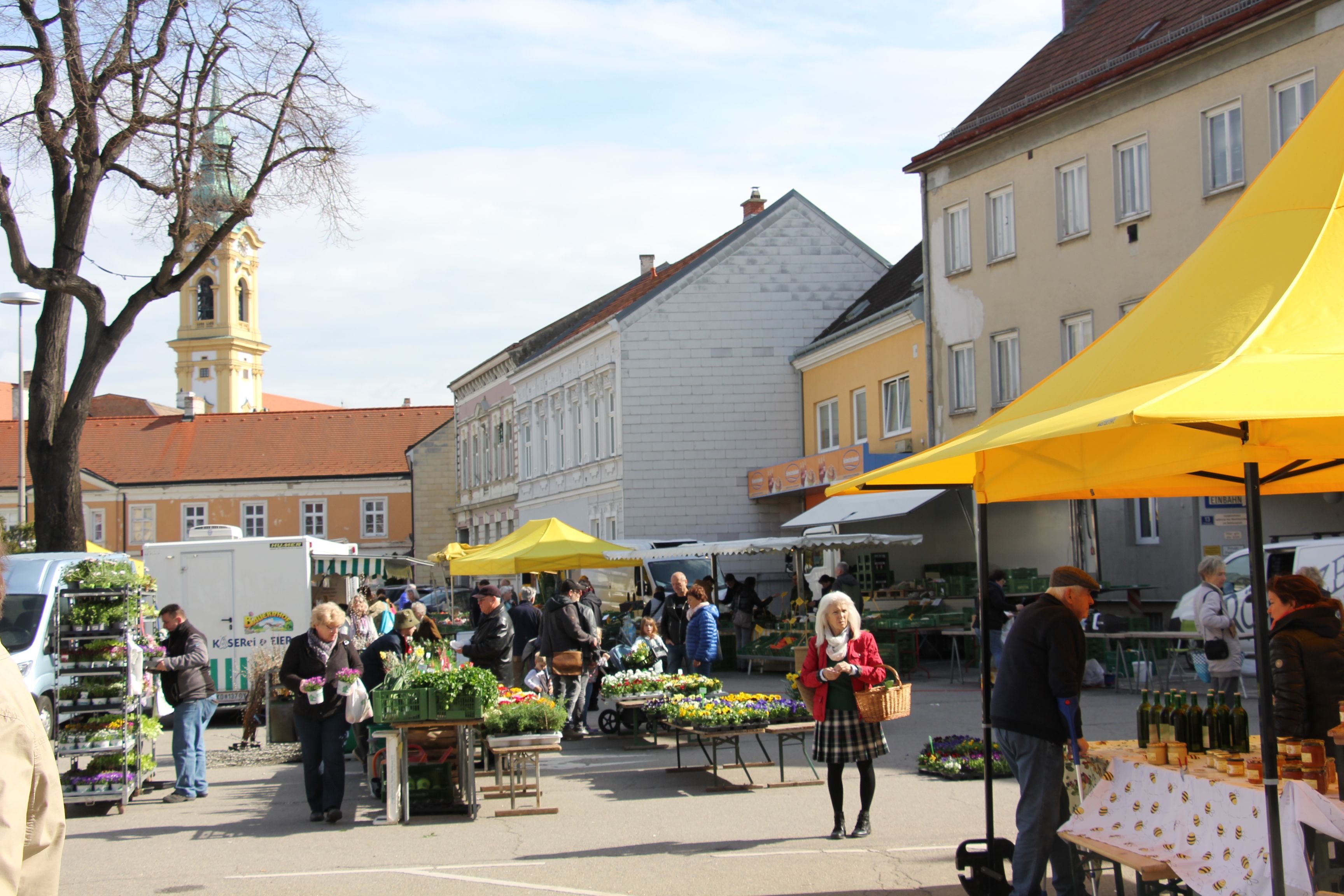 People at a weekly market in Stockerau with yellow market stalls and a church in the background.