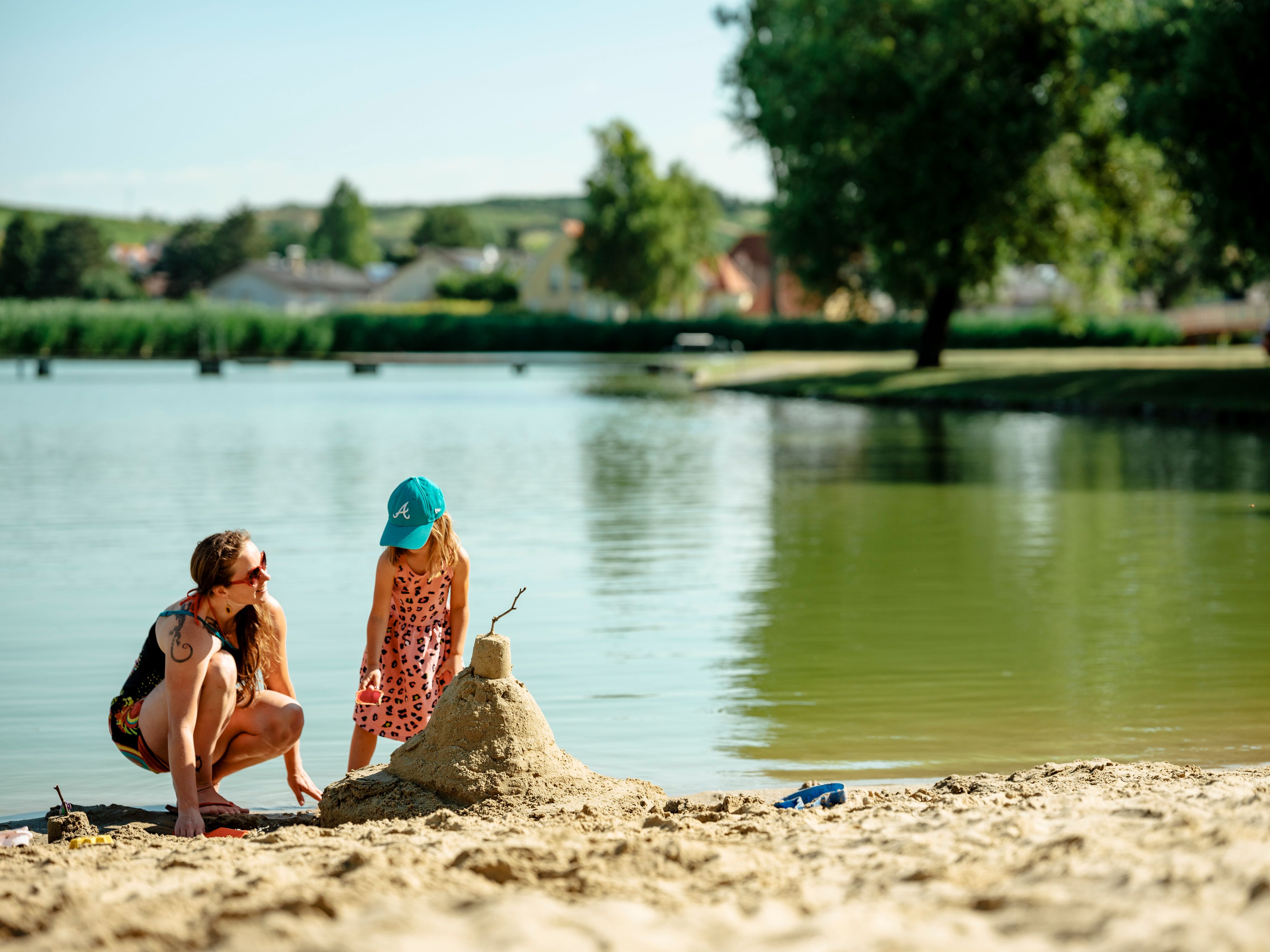 A woman and a child build a sandcastle on the shore of a swimming pond.