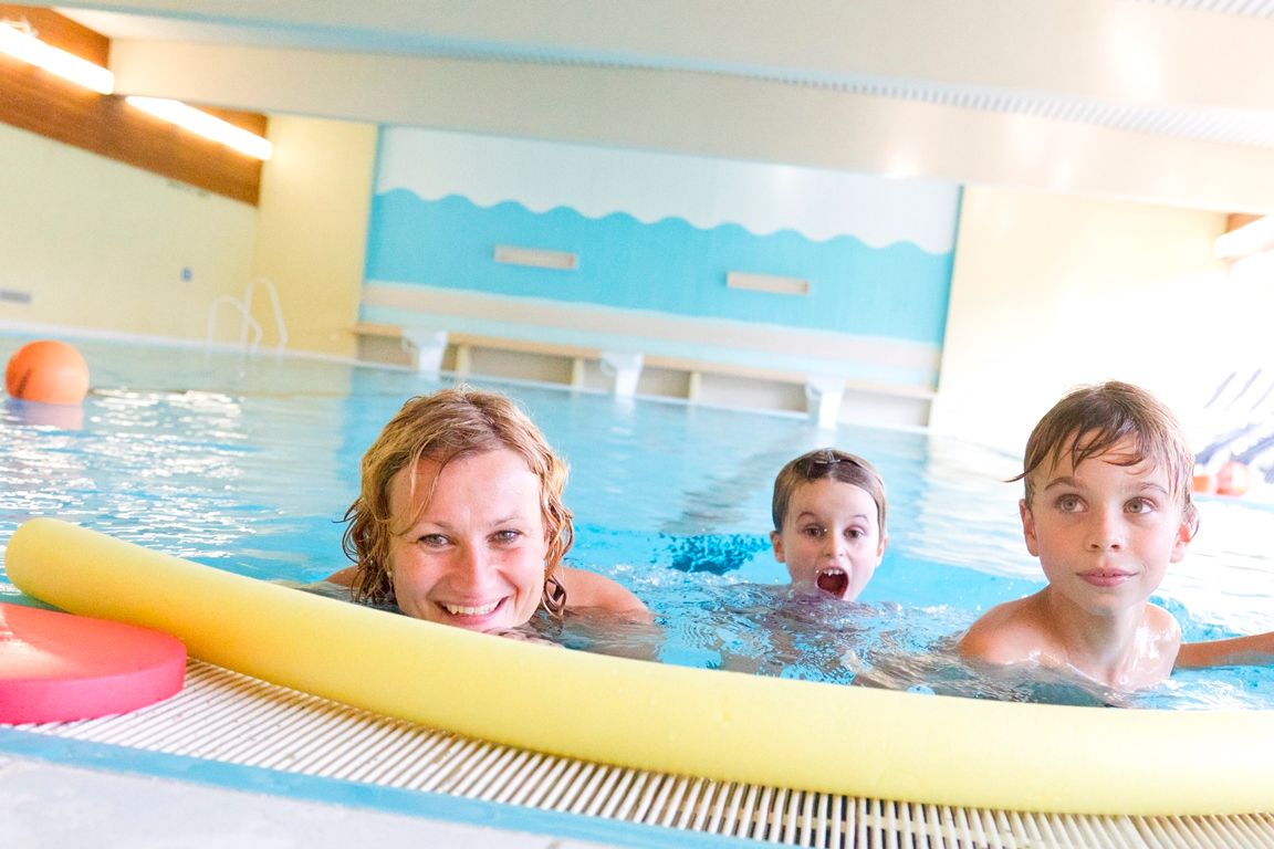 A woman and two children swim in an indoor pool, smiling and playing with a yellow pool noodle.