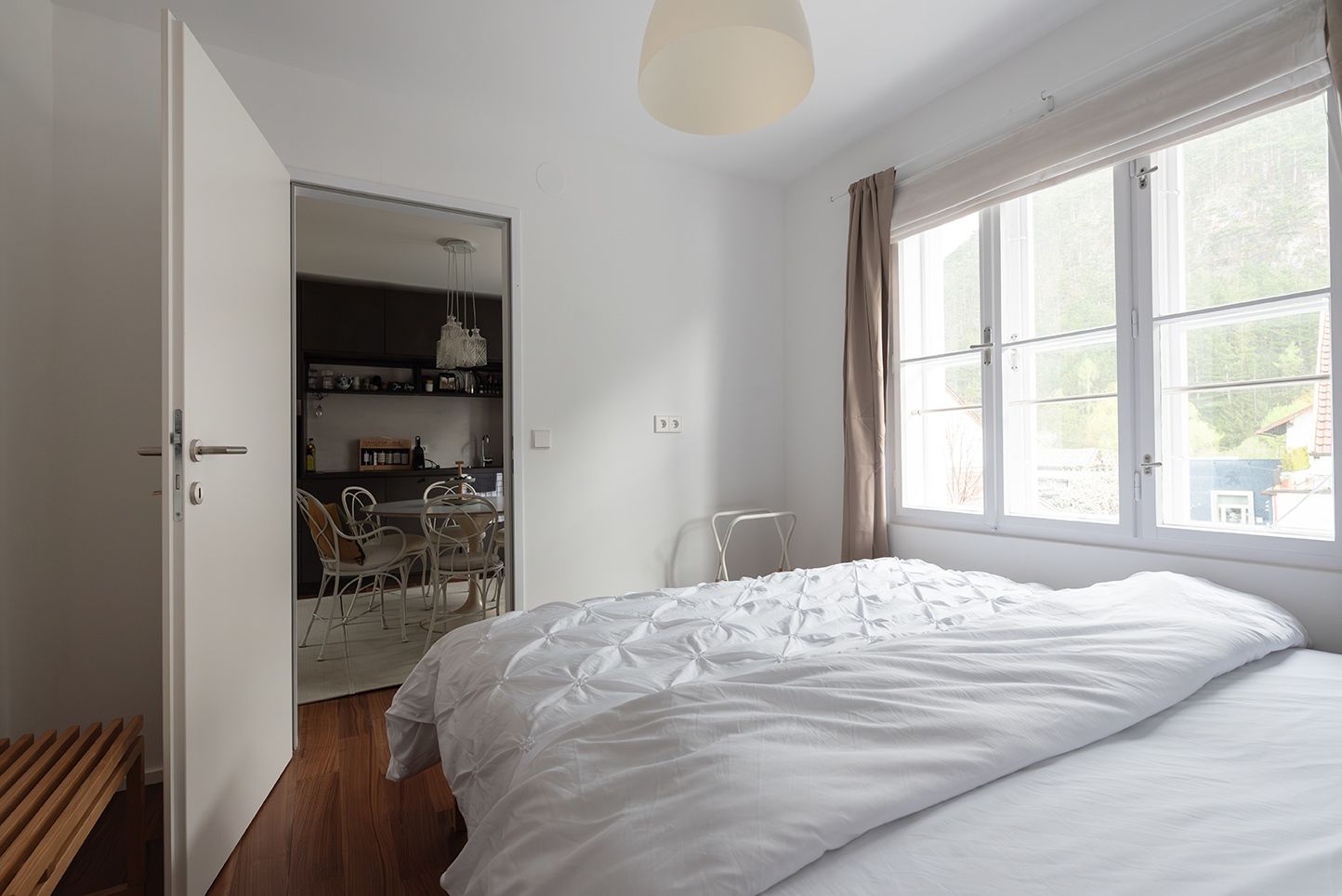 A bright bedroom with a large window, a bed and an open door leading to a dining area.
