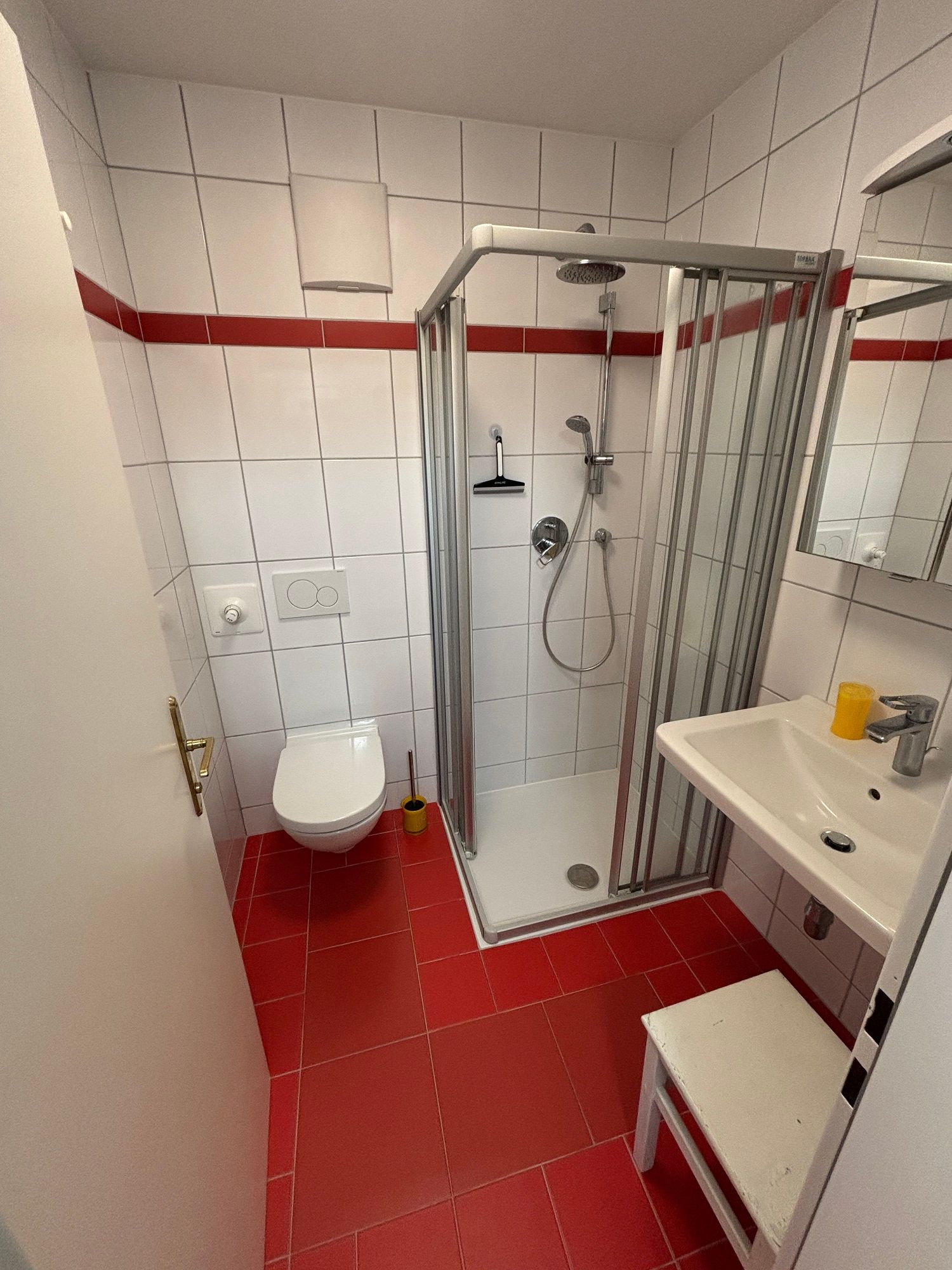Bathroom with red tiled floor, white tiled walls, shower, washbasin and toilet.