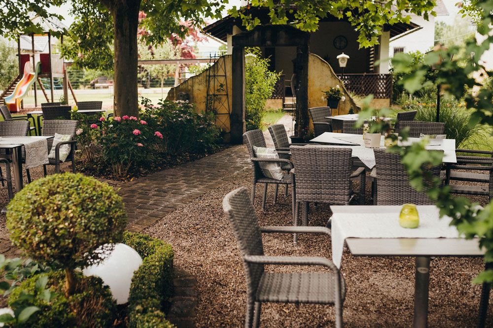 A cozy outdoor area with tables and chairs under trees, surrounded by plants and a playground in the background.