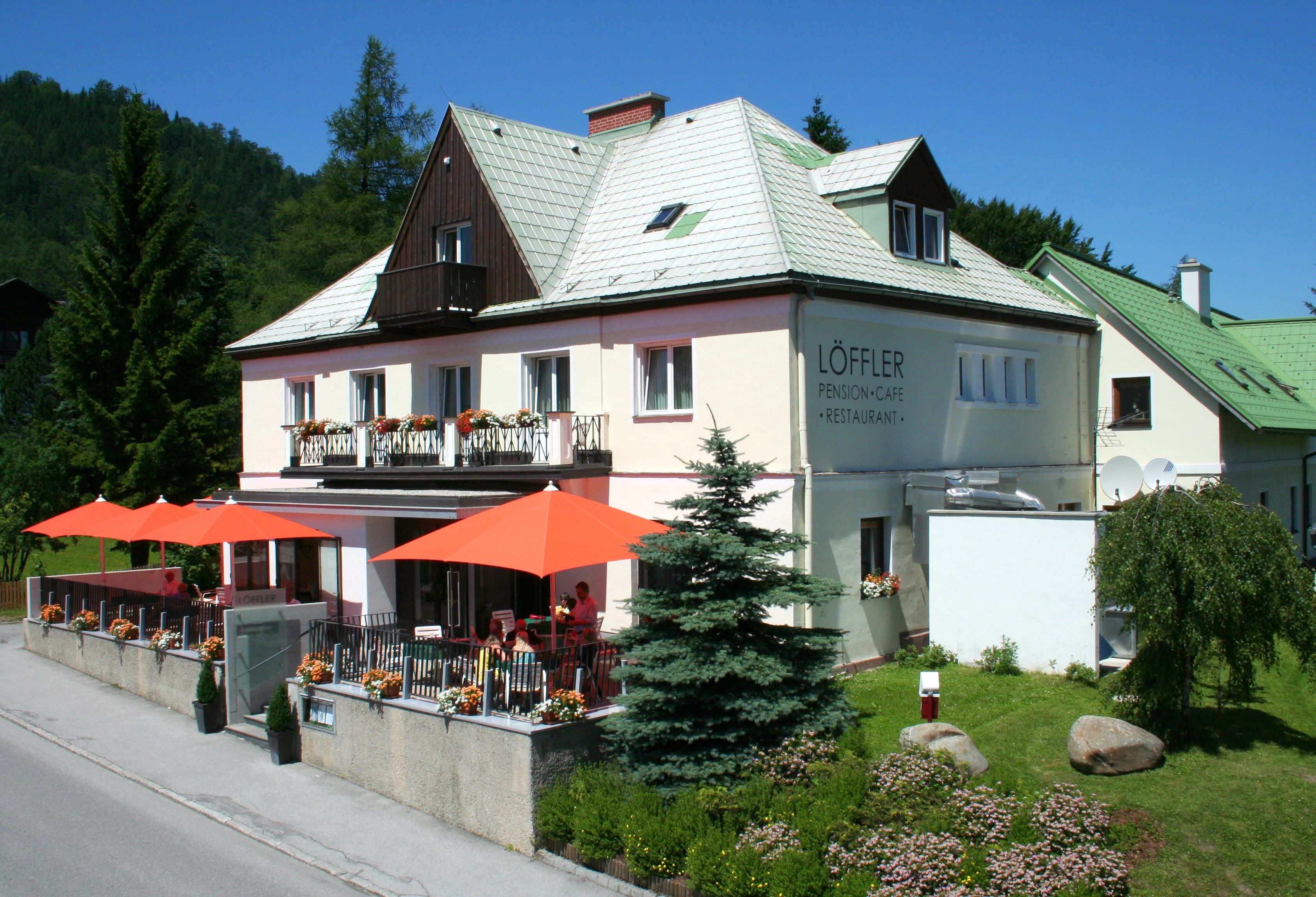 A two-storey building with the inscription 'Löffler Pension-Café-Restaurant', surrounded by trees and flowers, with orange parasols on the terrace.