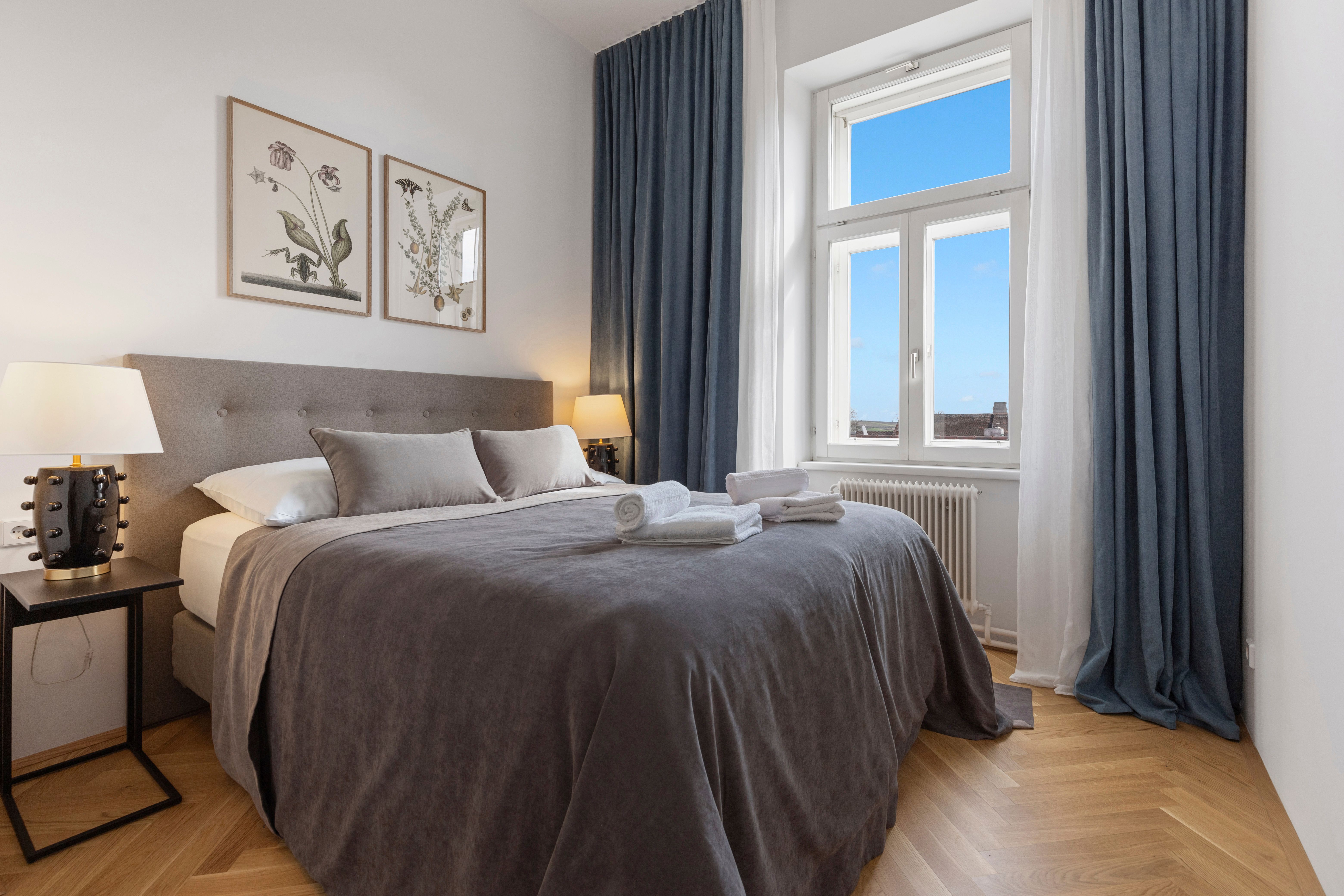 An elegant bedroom with a large bed, gray bed linen, two bedside tables with lamps and a window with a blue curtain.