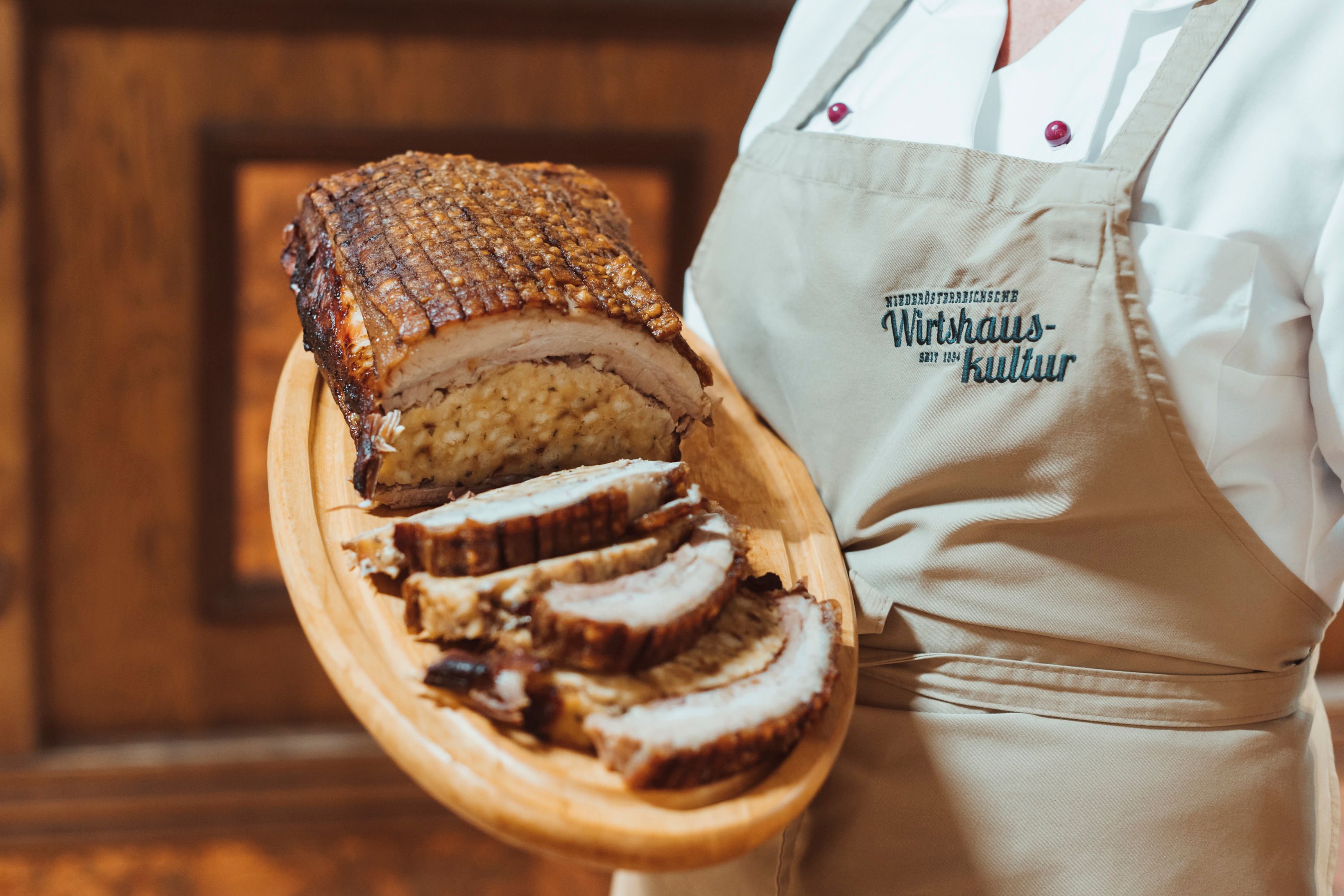 Stuffed pork breast on a wooden platter, served by a person in an apron.
