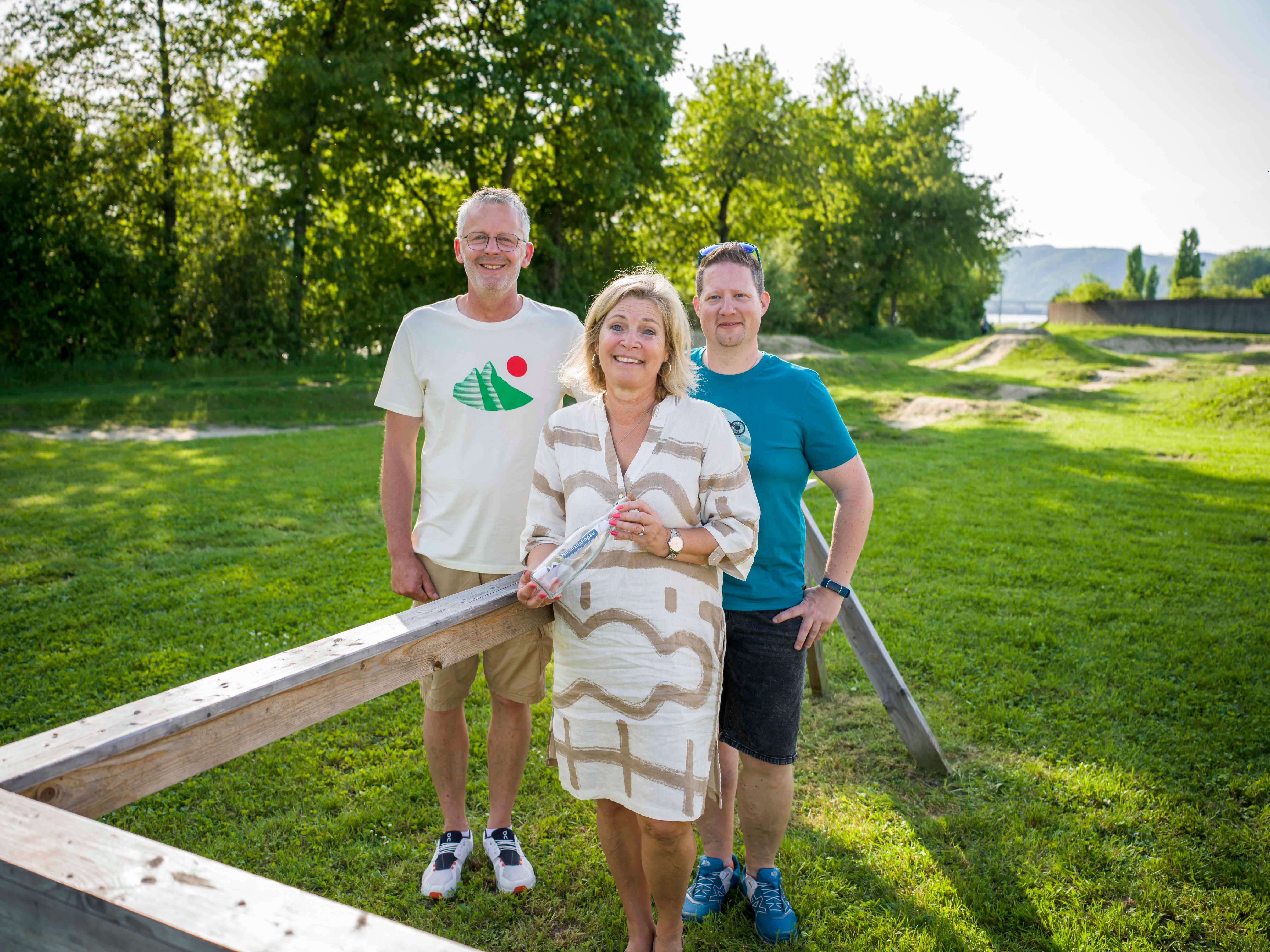 Three people are standing on a meadow in front of a wooden railing, surrounded by trees.