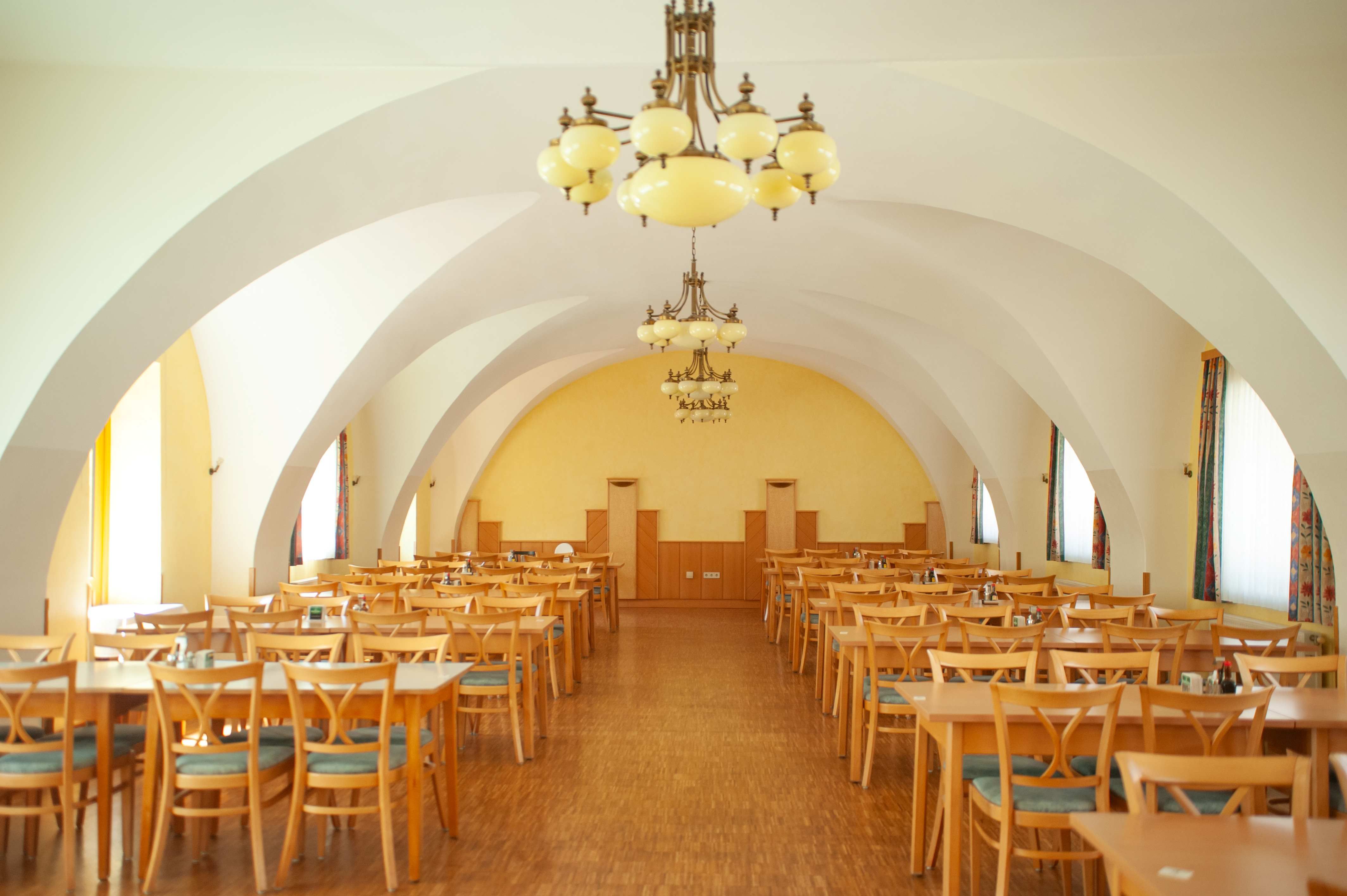 Interior view of a large dining room with wooden tables and chairs, yellow walls and decorative chandeliers.