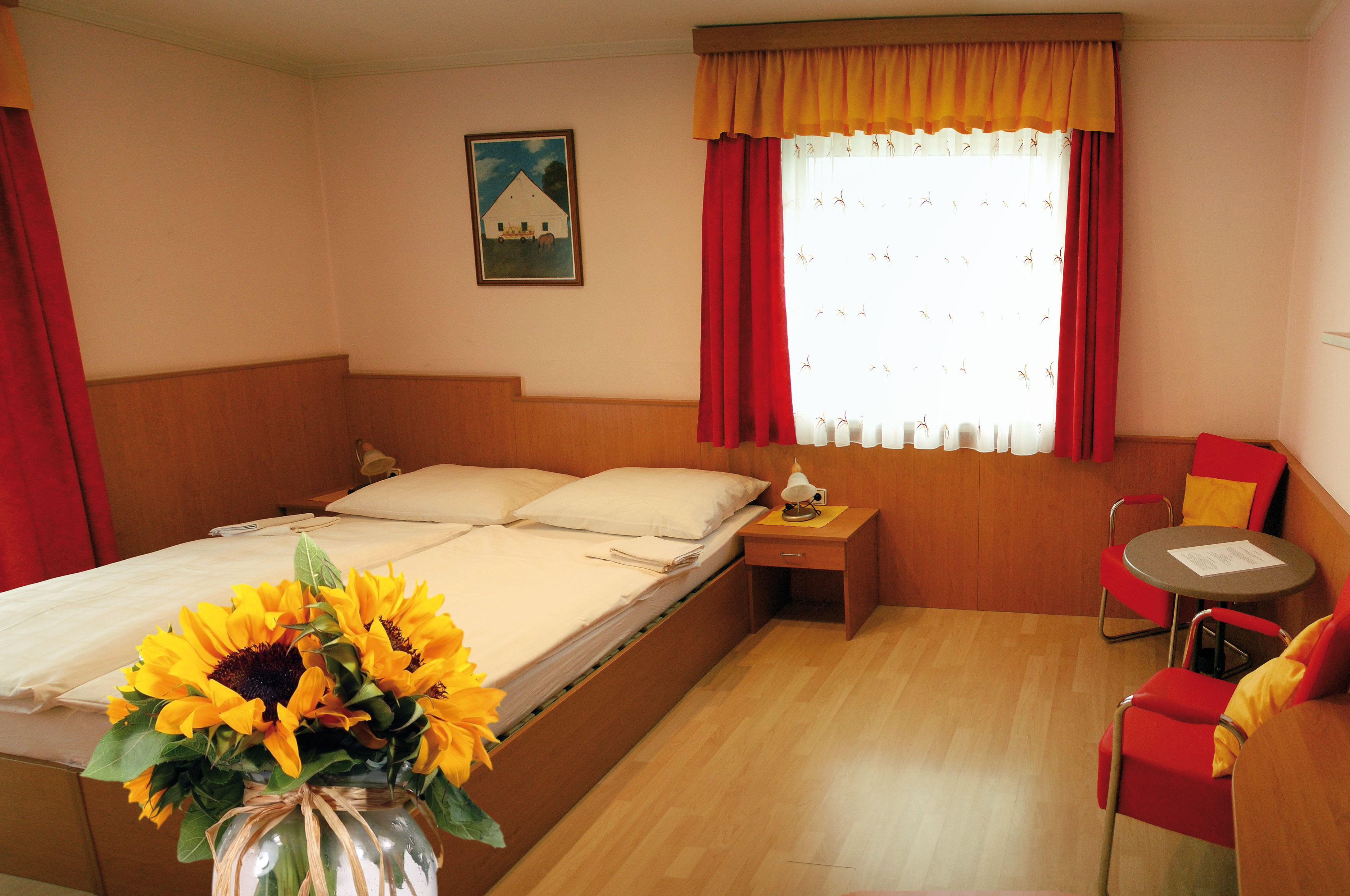 A cozy room with a double bed, red curtains, a table with chairs and a bouquet of sunflowers in the foreground.