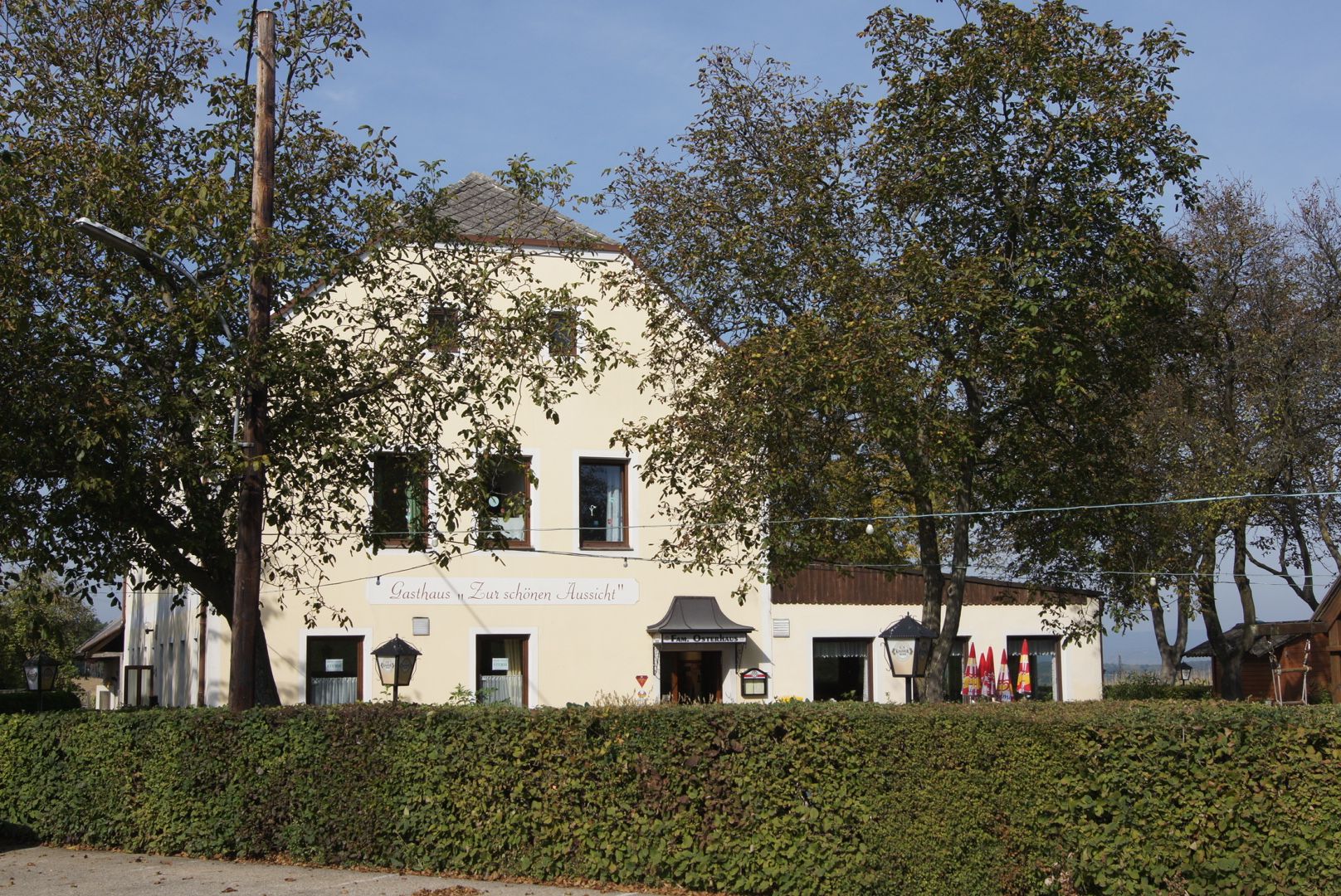 A yellow inn with the inscription 'Gasthaus Zur schönen Aussicht', surrounded by trees and hedges.