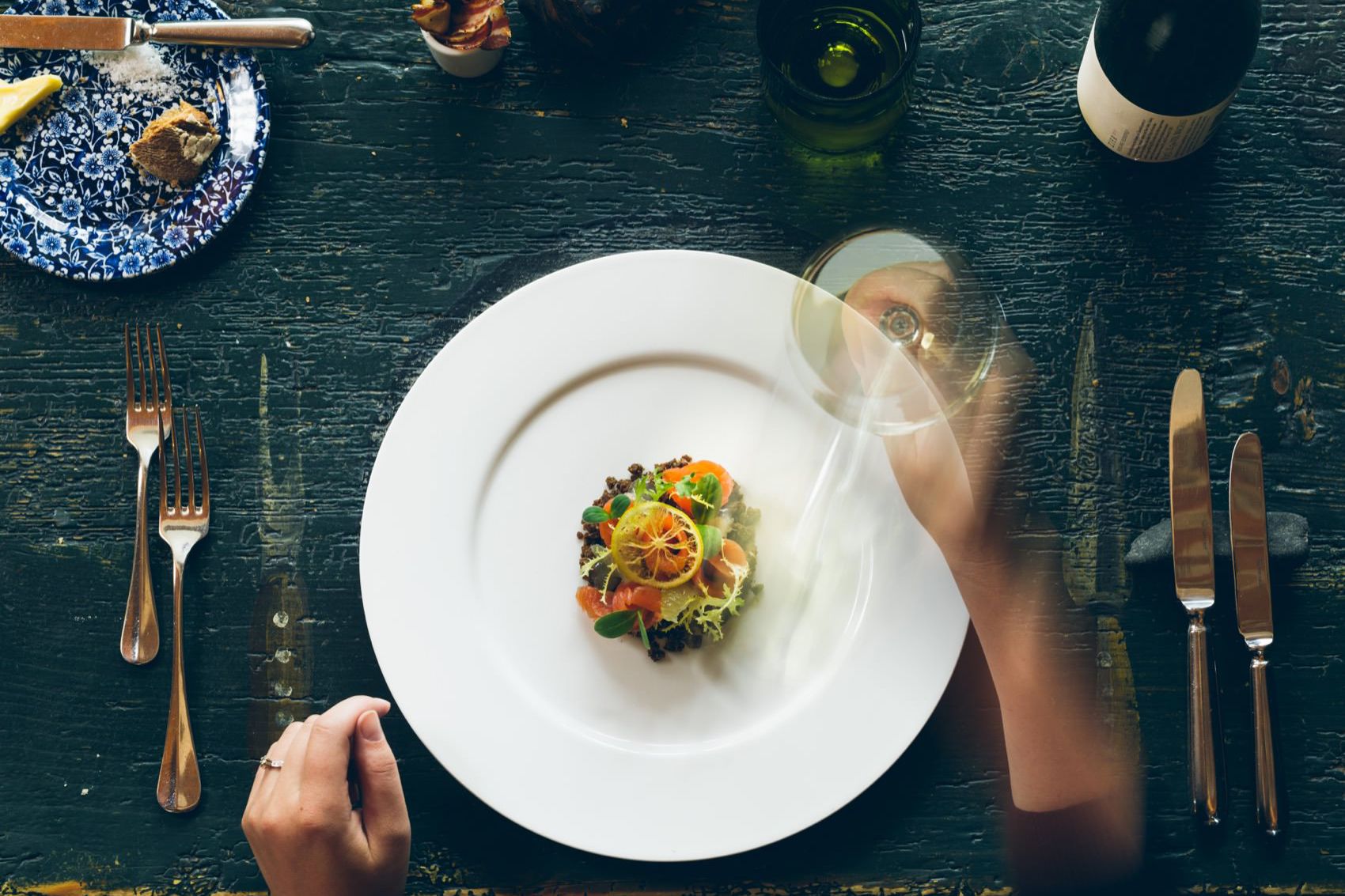 A plate with an artfully arranged dish, surrounded by cutlery and a wine glass on a rustic table.