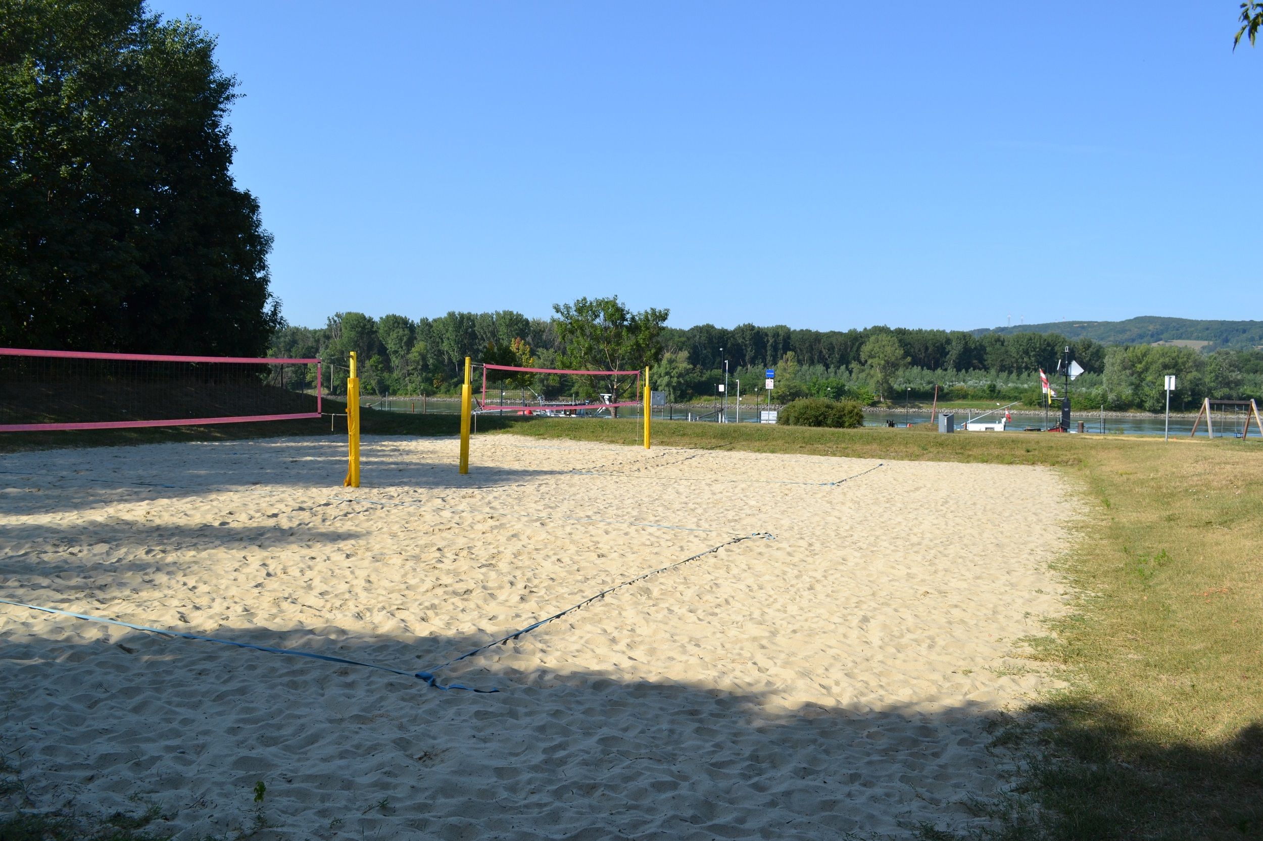 An empty beach volleyball court with yellow posts and a pink net, surrounded by trees and a river in the background.
