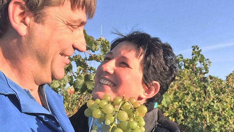A man and a woman smile at each other in a vineyard while holding a bunch of grapes.