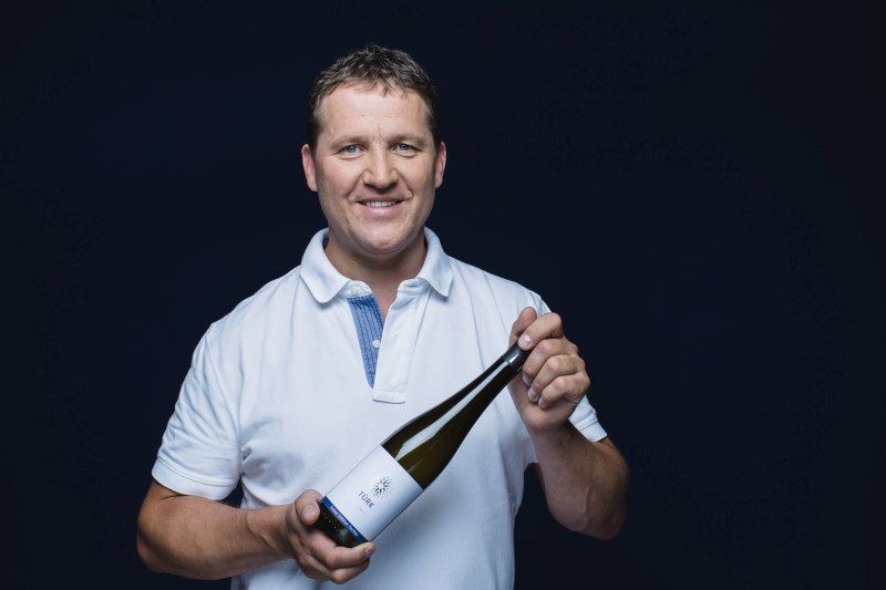 Man in white shirt holding a wine bottle with the label 'Türk'.