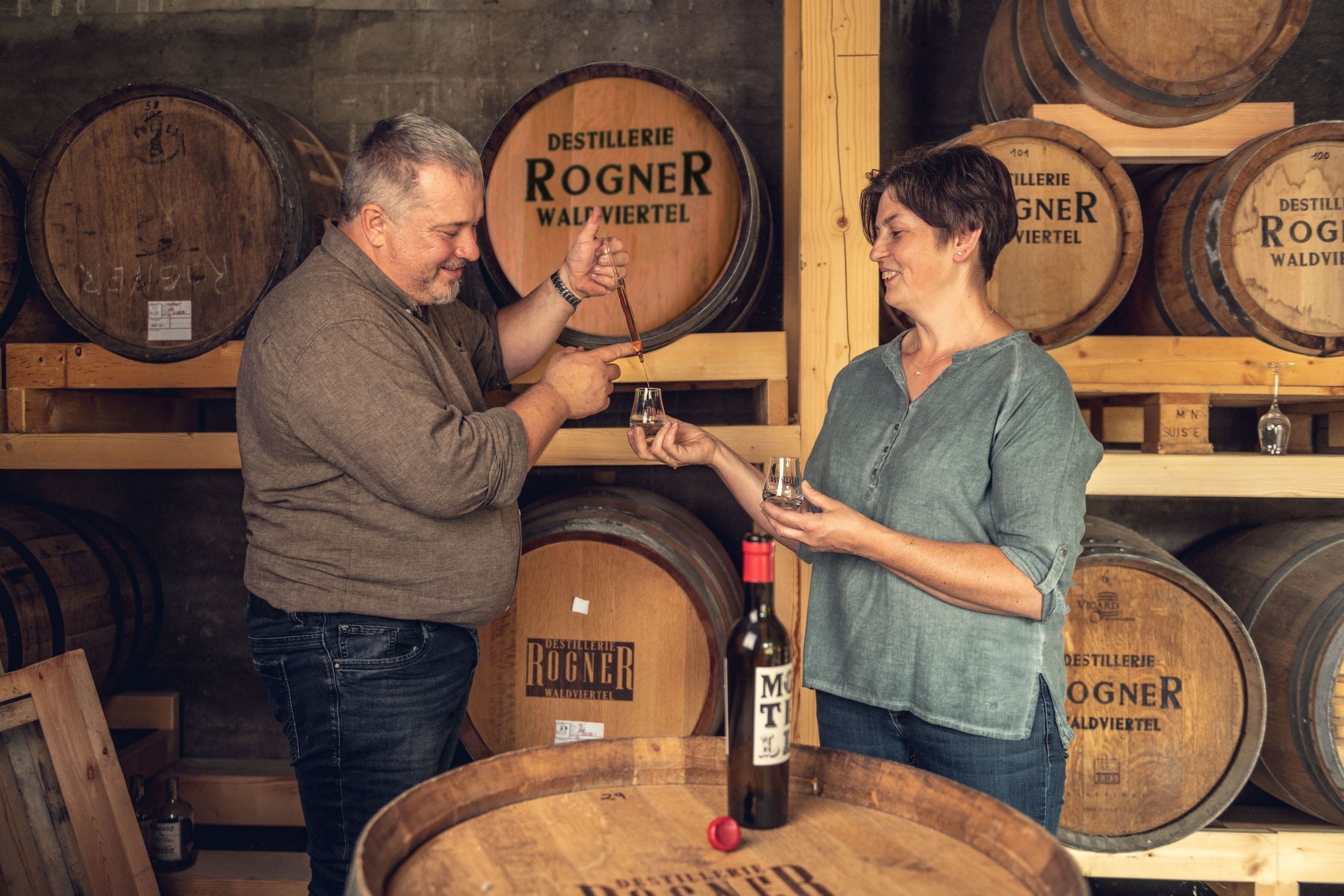 Two people at a tasting in a distillery with wooden barrels in the background.