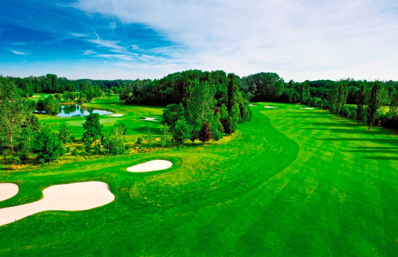 Green golf course with bunkers and pond, surrounded by trees under a blue sky.