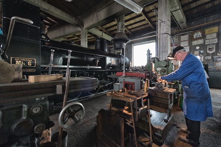 A man works in a workshop on a machine next to an old steam locomotive.