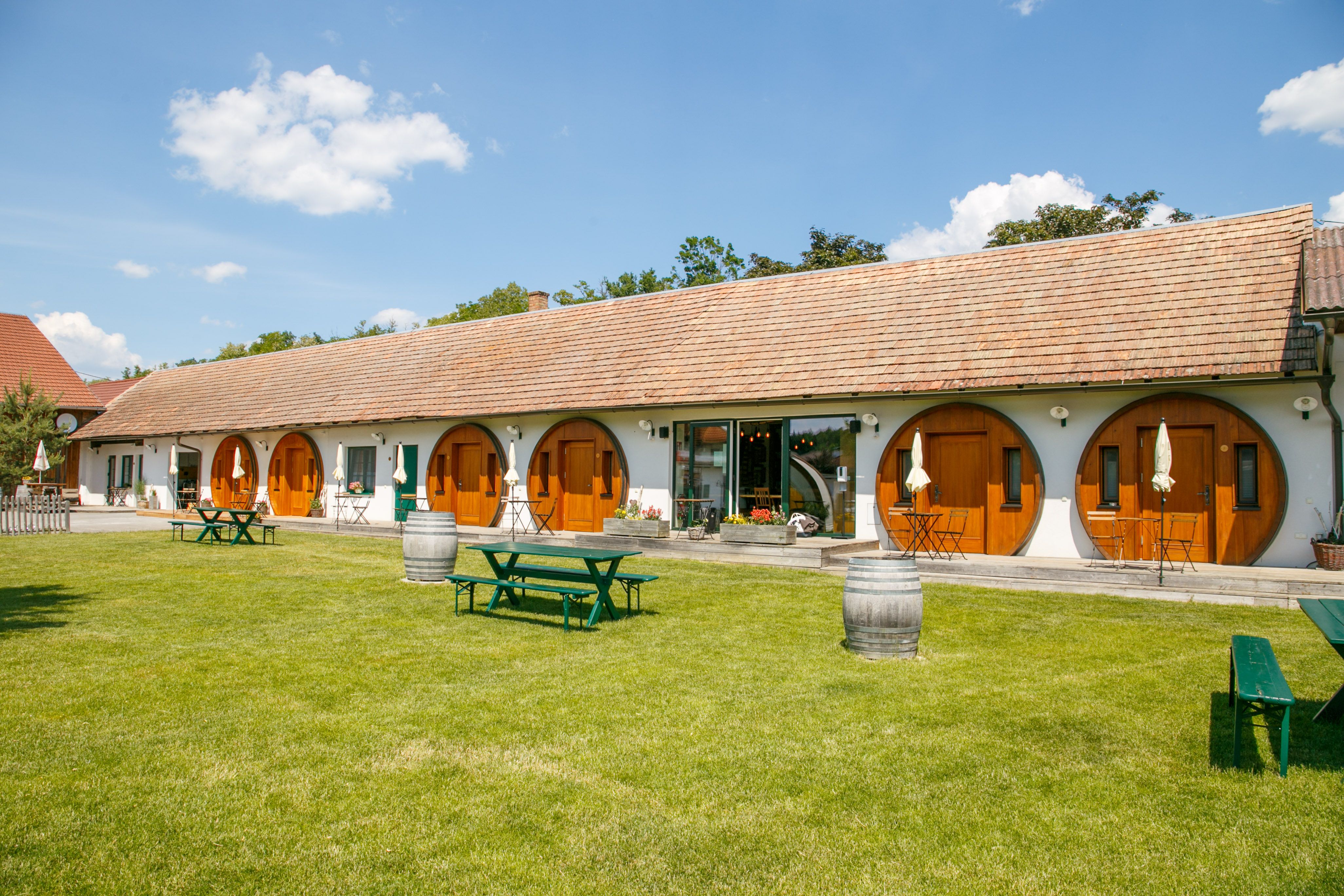 A long building with round wooden doors and a well-tended lawn in front of it with picnic tables and wine barrels.